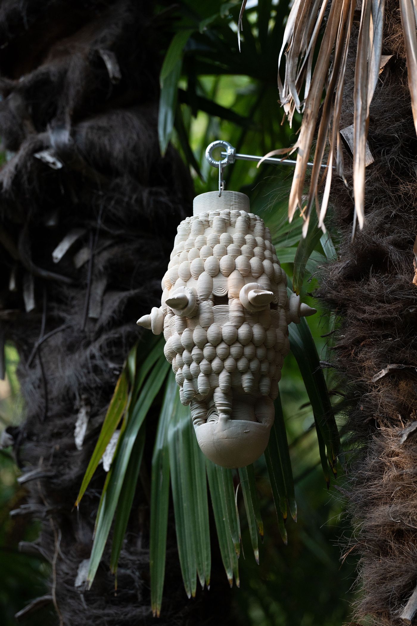 A unique, textured ceramic hanging art piece resembling a nest, suspended among lush palm leaves in a tropical setting. A unique, textured ceramic hanging art piece resembling a nest, suspended among lush palm leaves in a tropical setting.