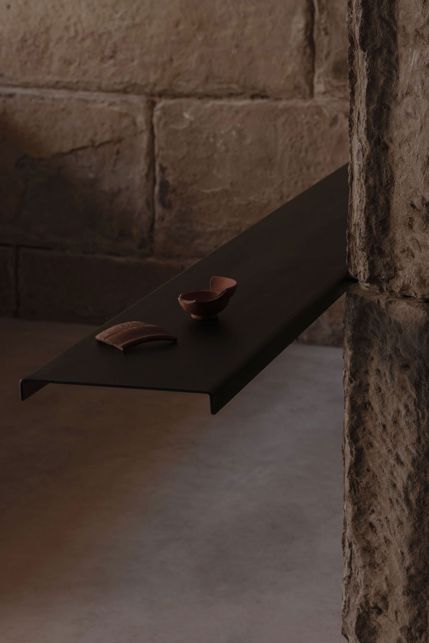 A sleek black shelf extends from a textured stone wall, holding two clay pottery pieces, illuminated softly in a minimalist space.