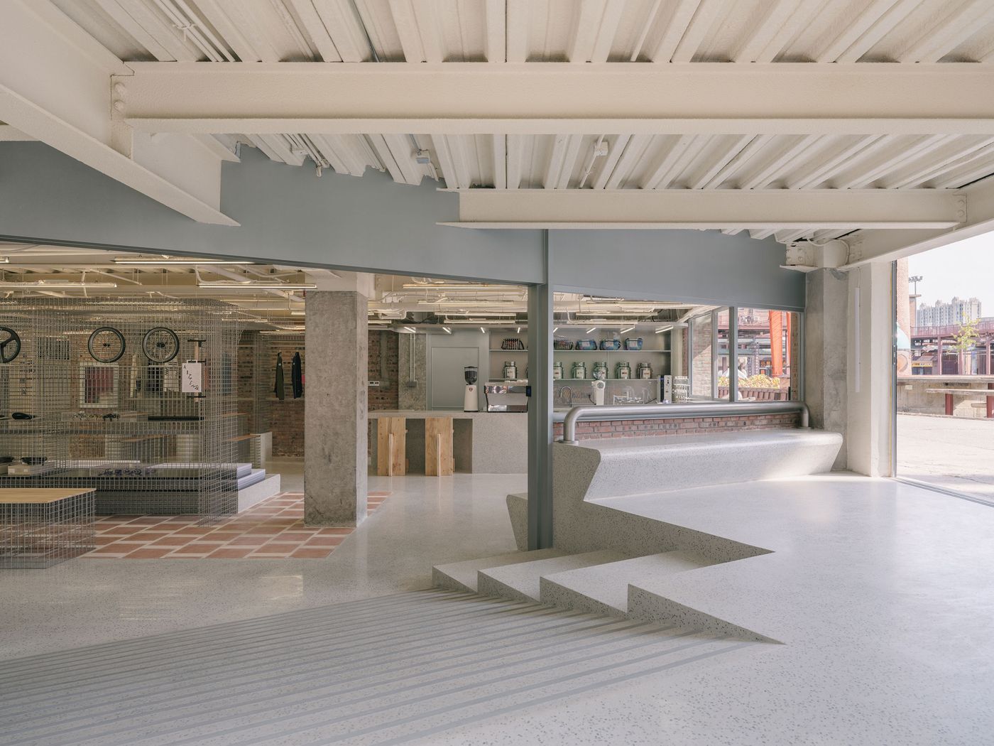 An expansive and brightly lit interior blends industrial robustness with a clean, contemporary feel. Exposed steel beams and a corrugated ceiling define the upper space, while polished concrete floors with geometric inlays lead to a striking stepped seating area. Beyond, a cafe counter and an open retail area with wire mesh displays suggest a multi-functional design. An expansive and brightly lit interior blends industrial robustness with a clean, contemporary feel. Exposed steel beams and a corrugated ceiling define the upper space, while polished concrete floors with geometric inlays lead to a striking stepped seating area. Beyond, a cafe counter and an open retail area with wire mesh displays suggest a multi-functional design.