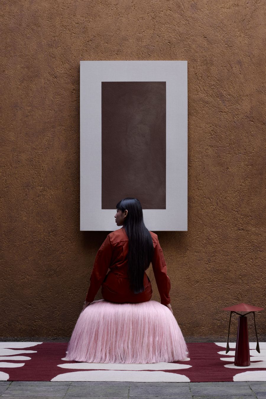 A person sits in a pink fringed skirt against a brown wall, gazing at a minimalist artwork above a patterned rug and modern stool.