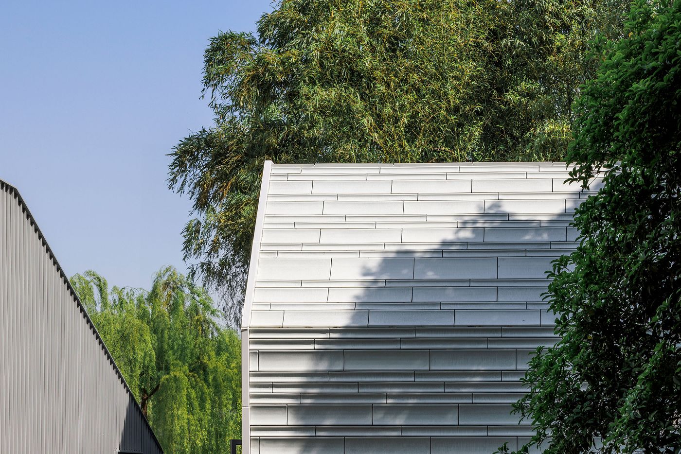 A compelling close-up reveals the textural interplay on the Lake House by Wutopia Lab. The primary building on the left features a vertical corrugated metal facade, while the adjacent section showcases a sophisticated, horizontal paneling in a lighter tone. Sunlight casts subtle shadows, highlighting the architectural details, with lush green foliage serving as a soft, natural backdrop, accentuating the contemporary design.