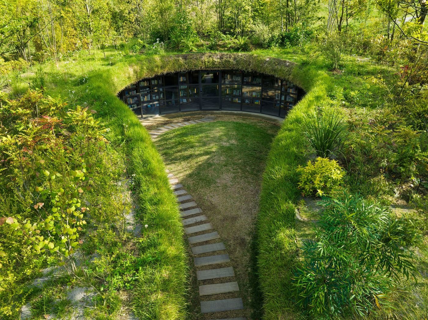A sunlit, circular library nestled in lush greenery, featuring a grass-covered roof and stone pathways leading into the garden. A sunlit, circular library nestled in lush greenery, featuring a grass-covered roof and stone pathways leading into the garden.