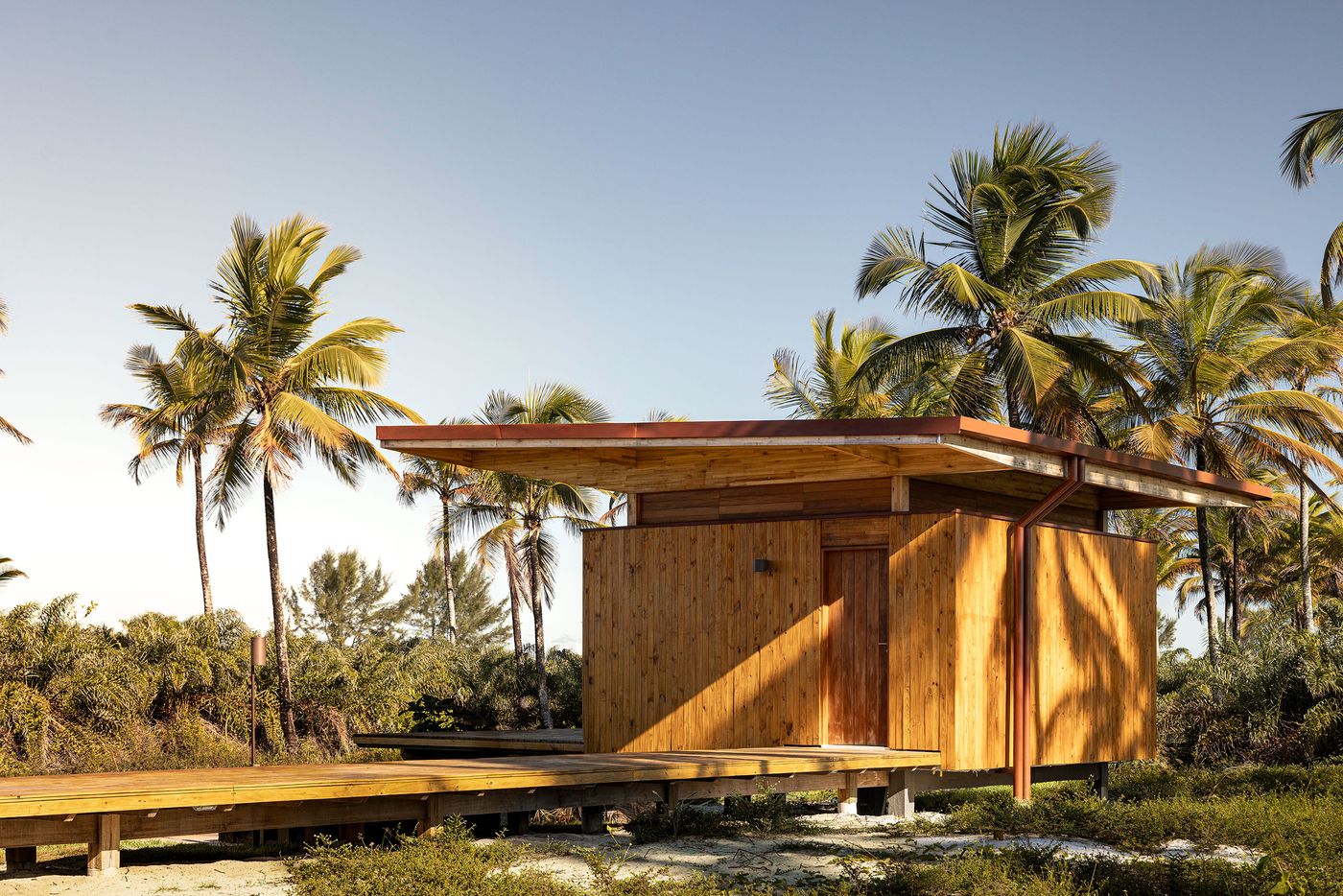 A wood-clad structure with a broad, gently sloping roof stands amidst a grove of palm trees. Elevated on a wooden deck, its minimalist form and natural materials blend effortlessly into the tropical environment, reflecting a biophilic and neo-vernacular design approach. A wood-clad structure with a broad, gently sloping roof stands amidst a grove of palm trees. Elevated on a wooden deck, its minimalist form and natural materials blend effortlessly into the tropical environment, reflecting a biophilic and neo-vernacular design approach.
