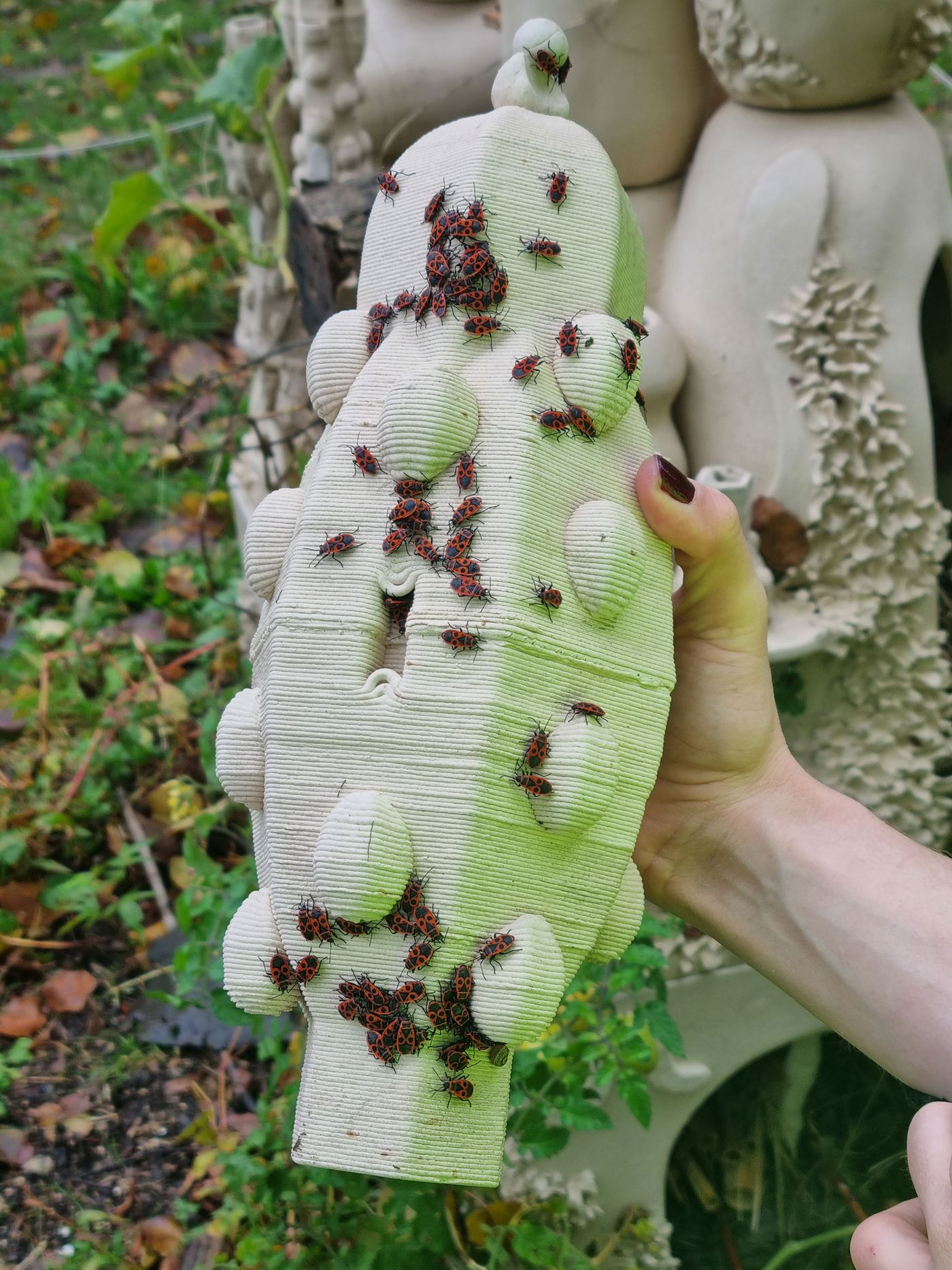 A hand holds a textured, cream-colored pottery sculpture covered with numerous red and black beetles in a lush garden setting. A hand holds a textured, cream-colored pottery sculpture covered with numerous red and black beetles in a lush garden setting.