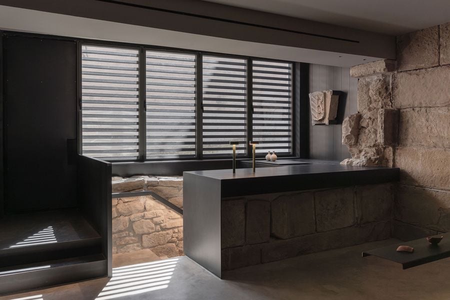 Modern interior featuring a black countertop, stone walls, and a large window with slatted blinds casting striped shadows.