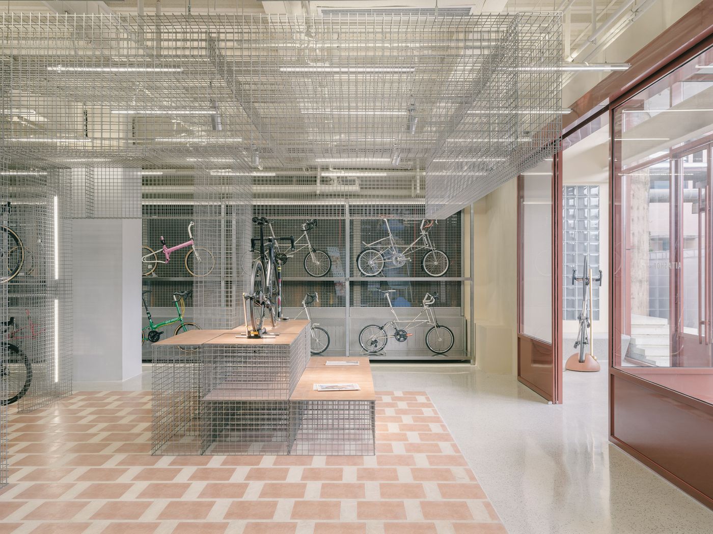 The interior offers a vast, uncluttered retail experience, emphasizing modular design with its ubiquitous wire mesh shelving and ceiling structures that elegantly display bicycles. A geometric pattern of terracotta-colored tiles delineates a central pathway, providing a warm textural contrast to the cool industrial materials and the polished concrete floor. The interior offers a vast, uncluttered retail experience, emphasizing modular design with its ubiquitous wire mesh shelving and ceiling structures that elegantly display bicycles. A geometric pattern of terracotta-colored tiles delineates a central pathway, providing a warm textural contrast to the cool industrial materials and the polished concrete floor.