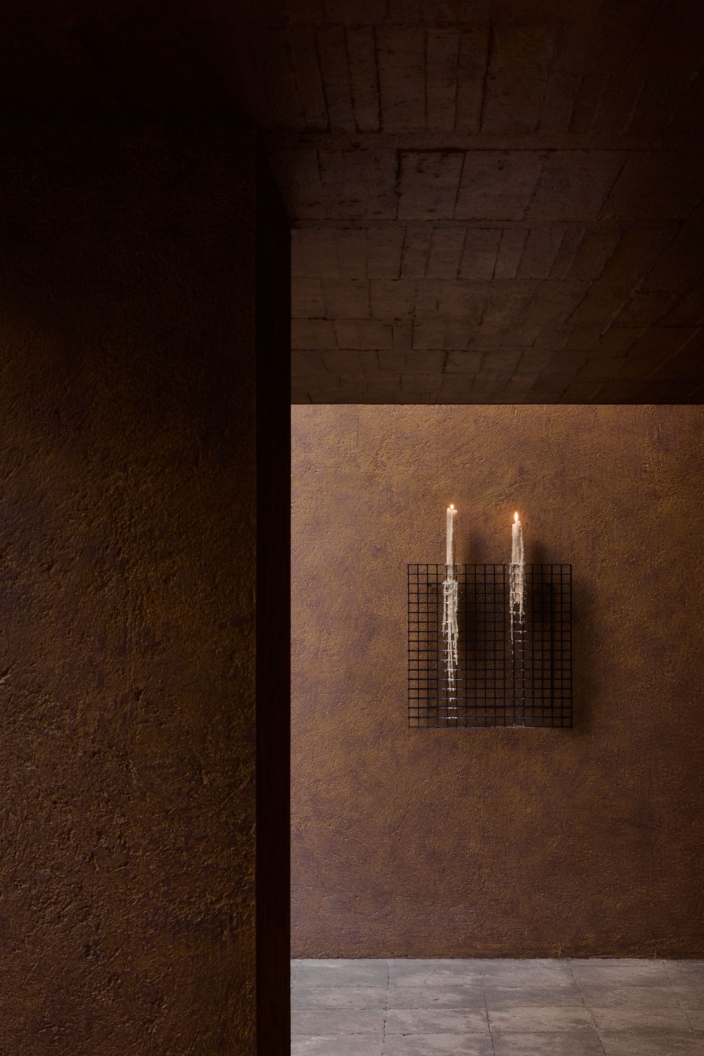 A dimly lit interior featuring a textured brown wall, with two elegant candles mounted in a black grid holder.