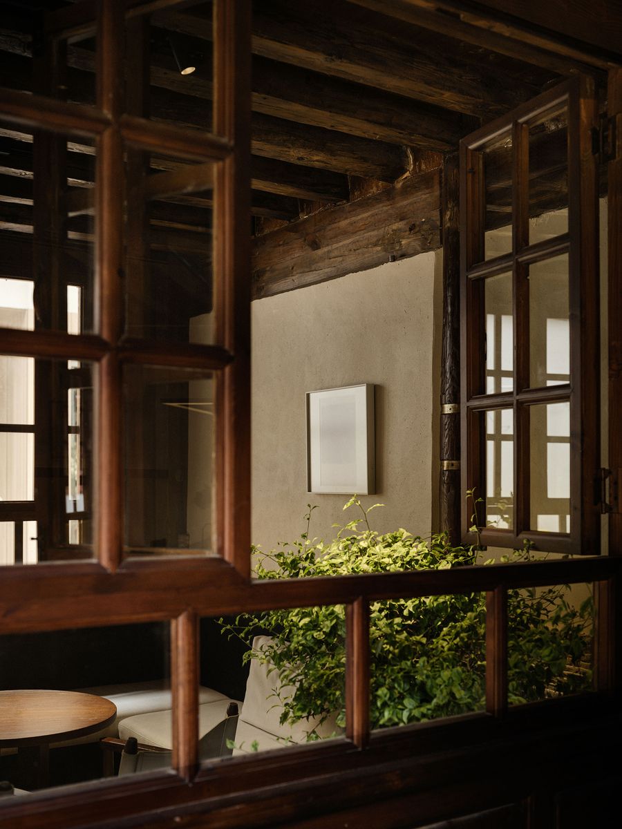 A cozy interior seen through wooden windows, featuring a small round table, a white sofa, art on the wall, and lush green plants. A cozy interior seen through wooden windows, featuring a small round table, a white sofa, art on the wall, and lush green plants.