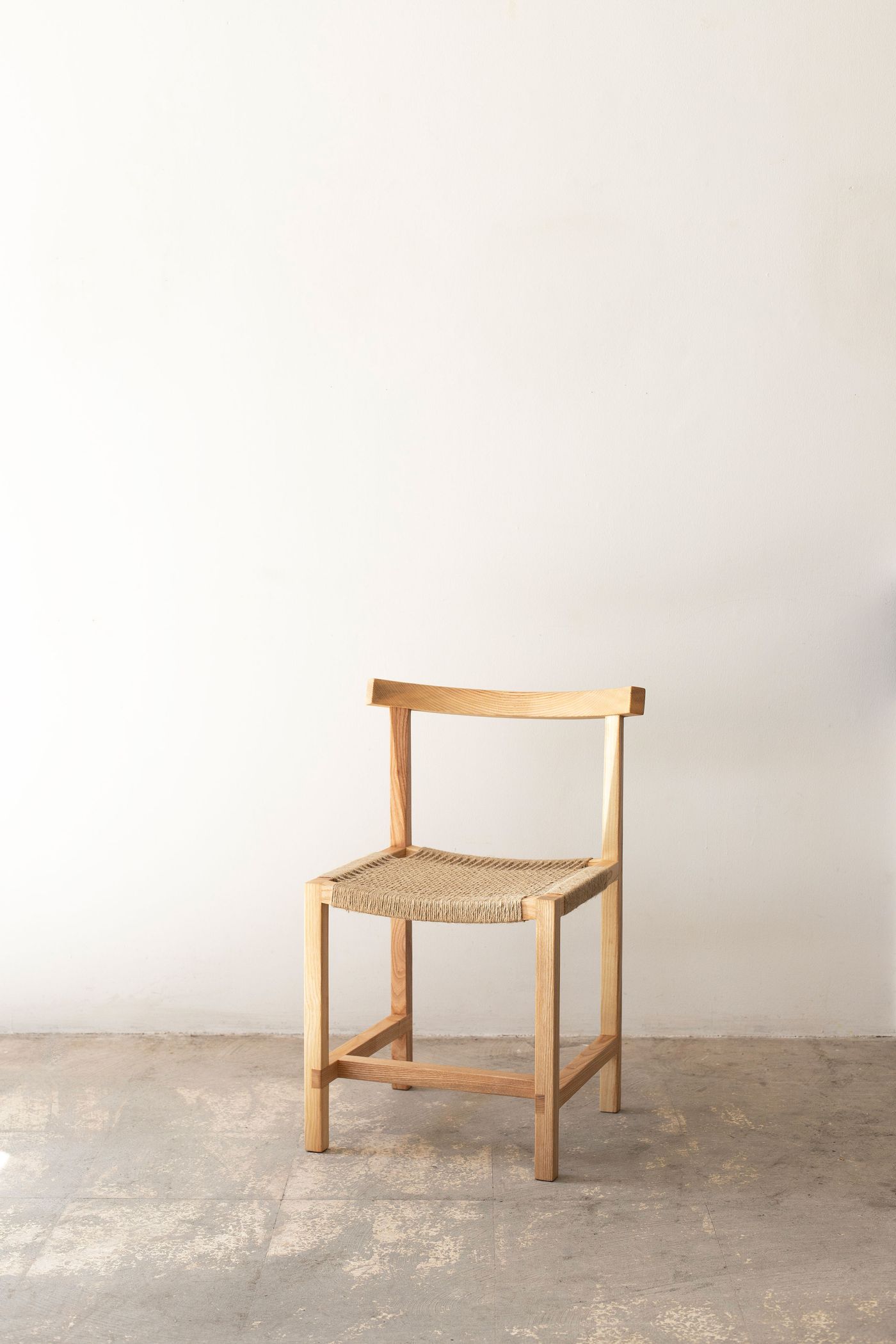 Pure Scandinavian restraint defines this simple dining chair with its clean ash frame and woven rush seat. The piece embodies functional minimalism, where every joint and angle serves both structural purpose and aesthetic harmony, standing confidently on weathered concrete flooring.