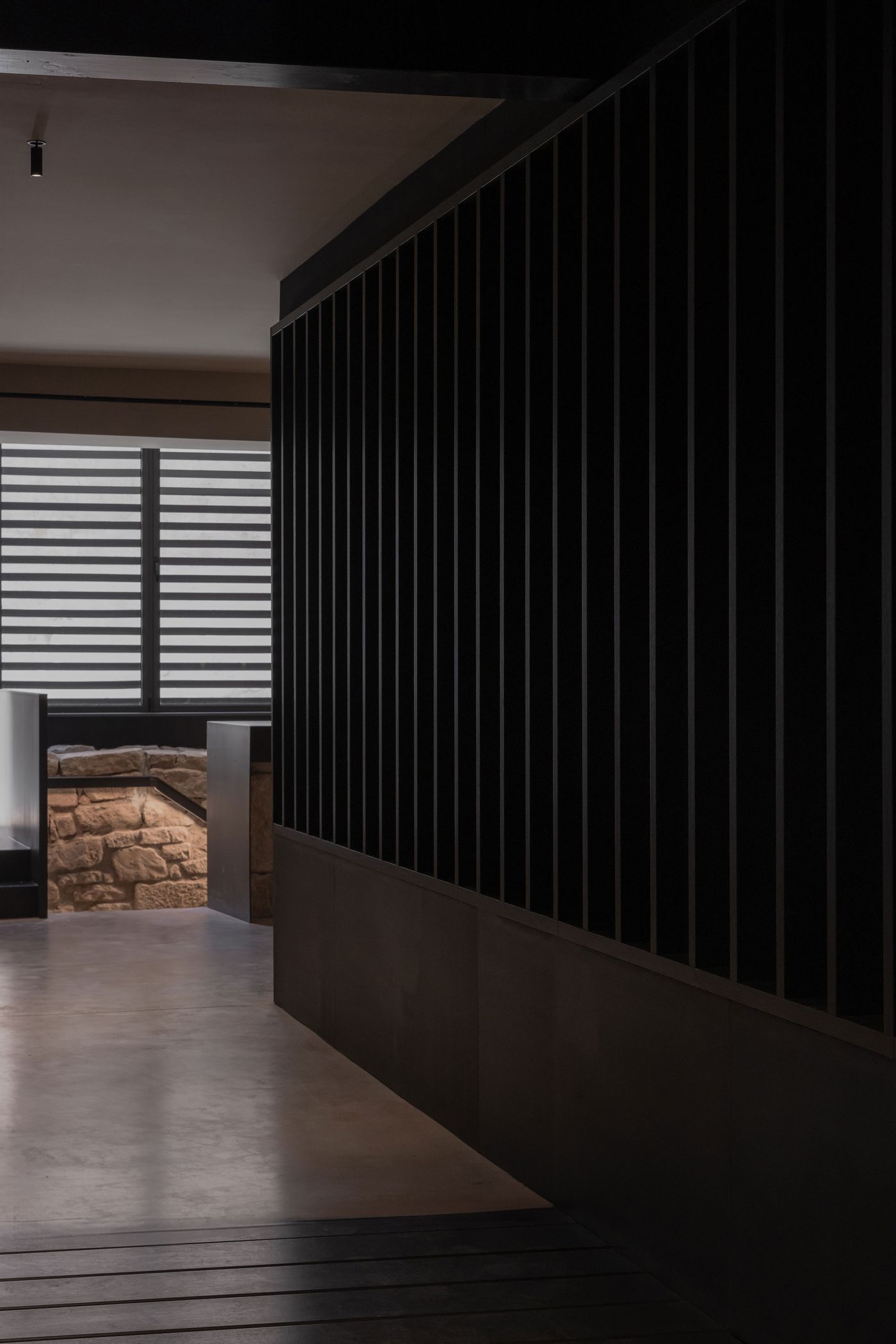 A dimly lit corridor with sleek black wooden slats on one wall, leading to a bright window with stone seating visible beyond.