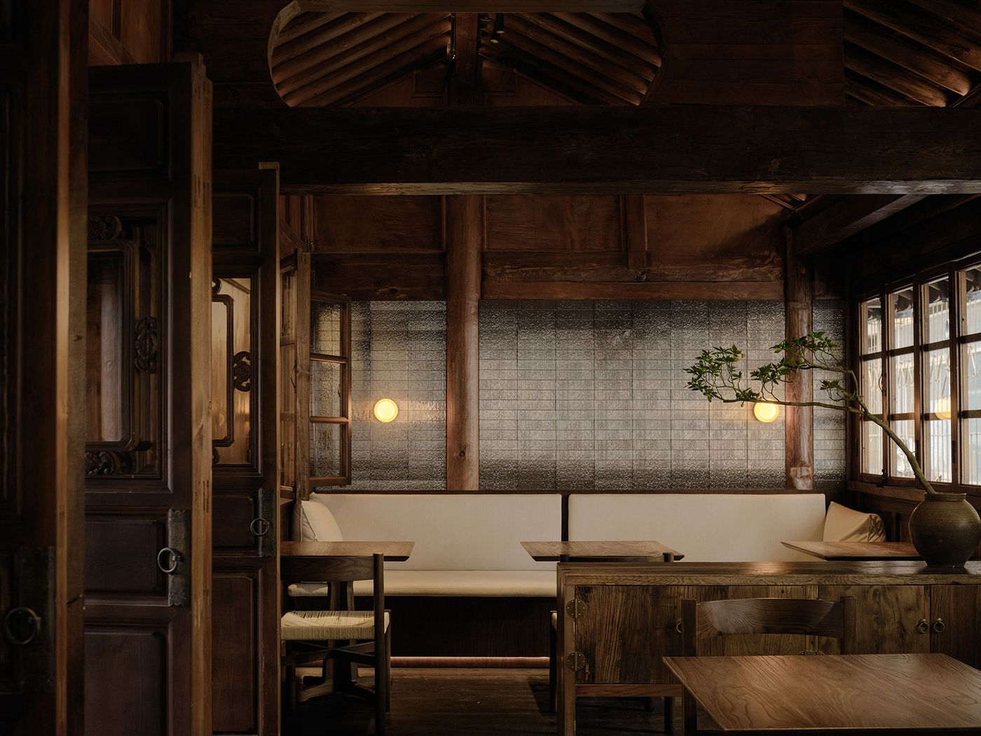 Cozy wooden interior of a restaurant featuring warm lighting, a simple table, plush seating, and a decorative plant in a vase. Cozy wooden interior of a restaurant featuring warm lighting, a simple table, plush seating, and a decorative plant in a vase.