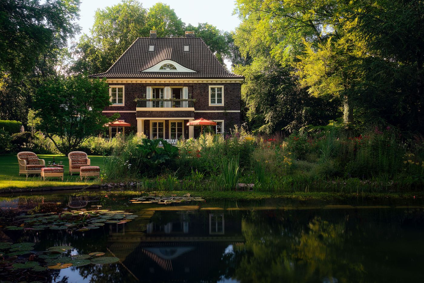 A beautiful, large house with a balcony, surrounded by lush greenery and a serene pond, featuring cozy chairs and colorful flowers. A beautiful, large house with a balcony, surrounded by lush greenery and a serene pond, featuring cozy chairs and colorful flowers.