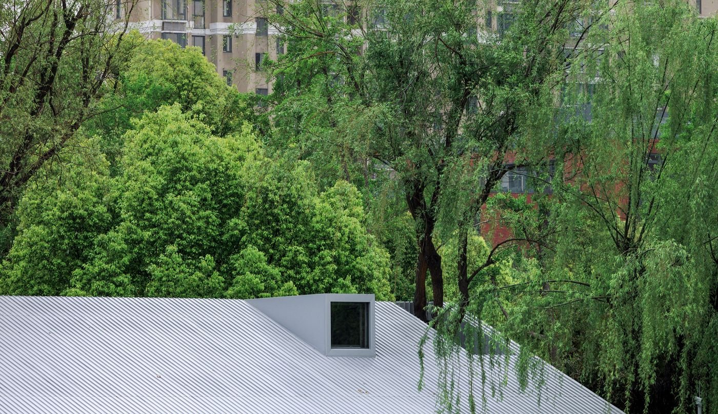 A close-up of the Lake House by Wutopia Lab reveals its crisp, gabled roof clad in shimmering, light-grey corrugated metal, punctuated by a dark, minimalist dormer window. Verdant green trees, including a weeping willow, frame the structure against a soft sky, subtly contrasting the modern, precise architecture with the organic forms of nature and distant urban buildings. A close-up of the Lake House by Wutopia Lab reveals its crisp, gabled roof clad in shimmering, light-grey corrugated metal, punctuated by a dark, minimalist dormer window. Verdant green trees, including a weeping willow, frame the structure against a soft sky, subtly contrasting the modern, precise architecture with the organic forms of nature and distant urban buildings.