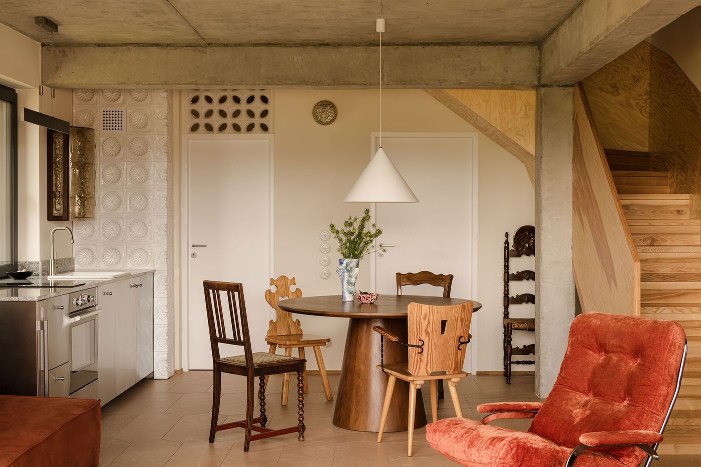 A modern dining area featuring a round wooden table, eclectic chairs, a vase of flowers, and a wooden staircase in a warm-toned space. A modern dining area featuring a round wooden table, eclectic chairs, a vase of flowers, and a wooden staircase in a warm-toned space.