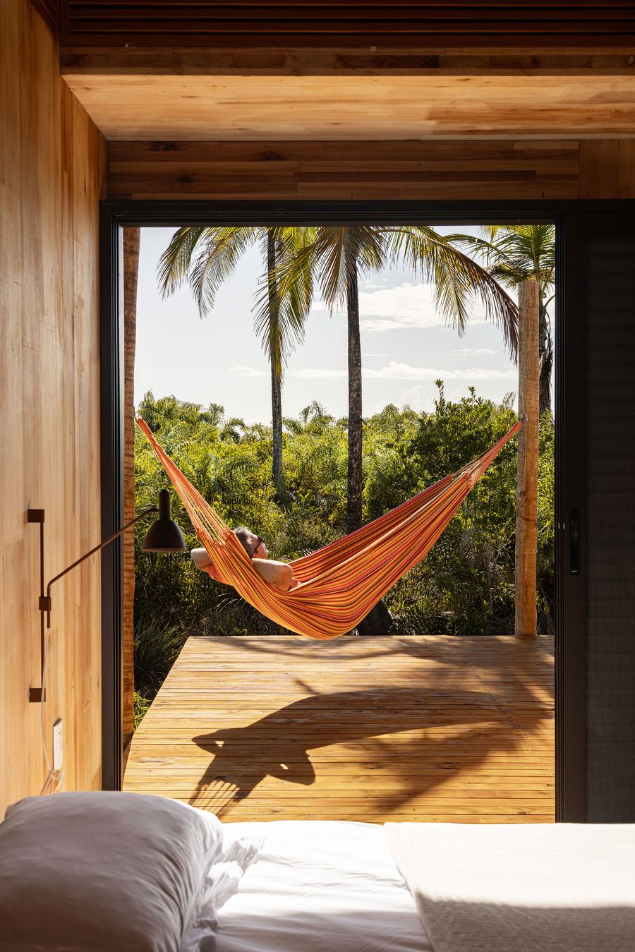 A person relaxes in a vibrant striped hammock, suspended on a wooden deck with a backdrop of lush tropical foliage and palm trees. The view from inside a simple, wood-paneled room emphasizes a fluid indoor-outdoor connection, evoking a sense of tranquil escapism. A person relaxes in a vibrant striped hammock, suspended on a wooden deck with a backdrop of lush tropical foliage and palm trees. The view from inside a simple, wood-paneled room emphasizes a fluid indoor-outdoor connection, evoking a sense of tranquil escapism.