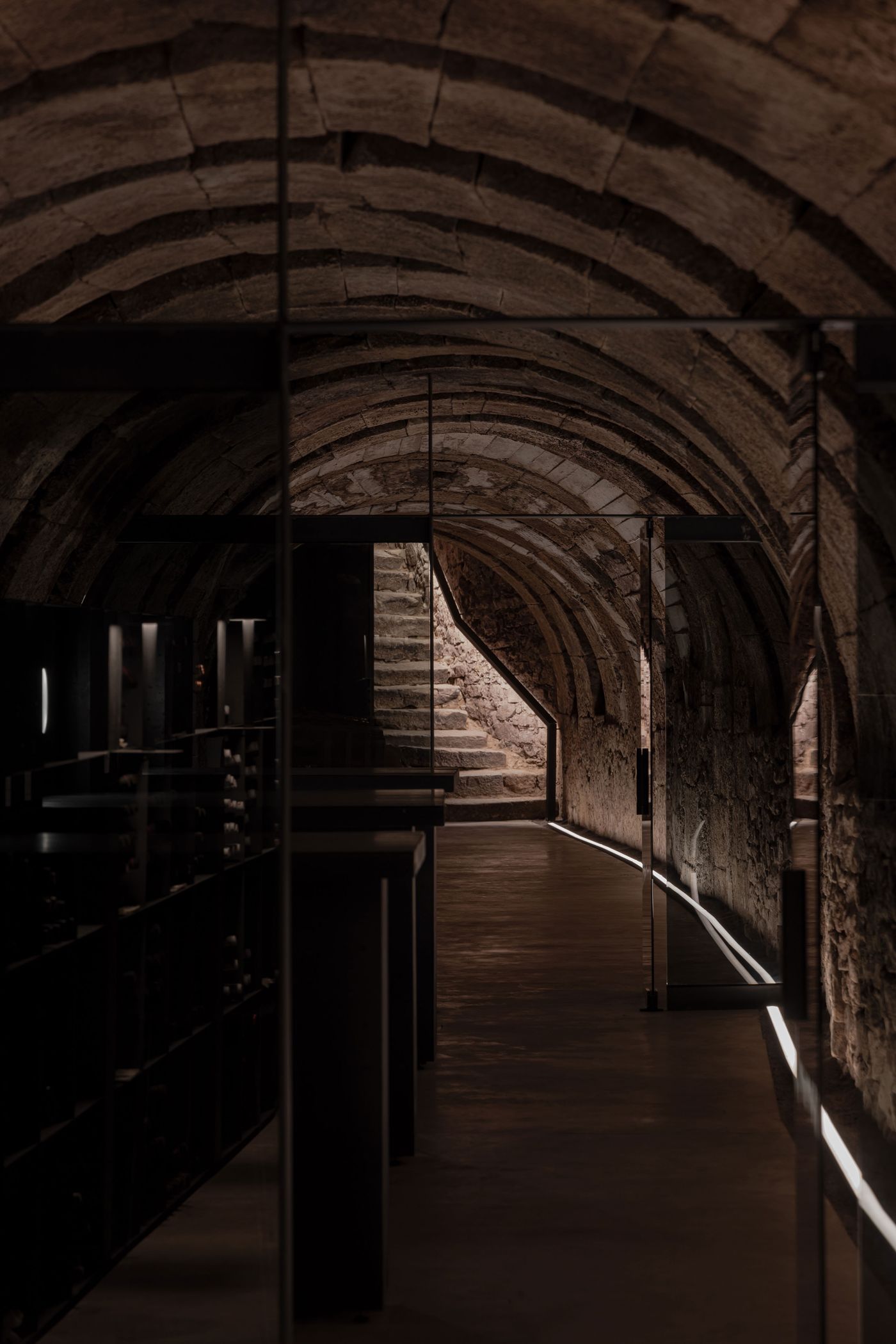 A dimly lit stone cellar with arched ceilings, showcasing a curved path and lit staircase leading upward, flanked by wine storage racks.