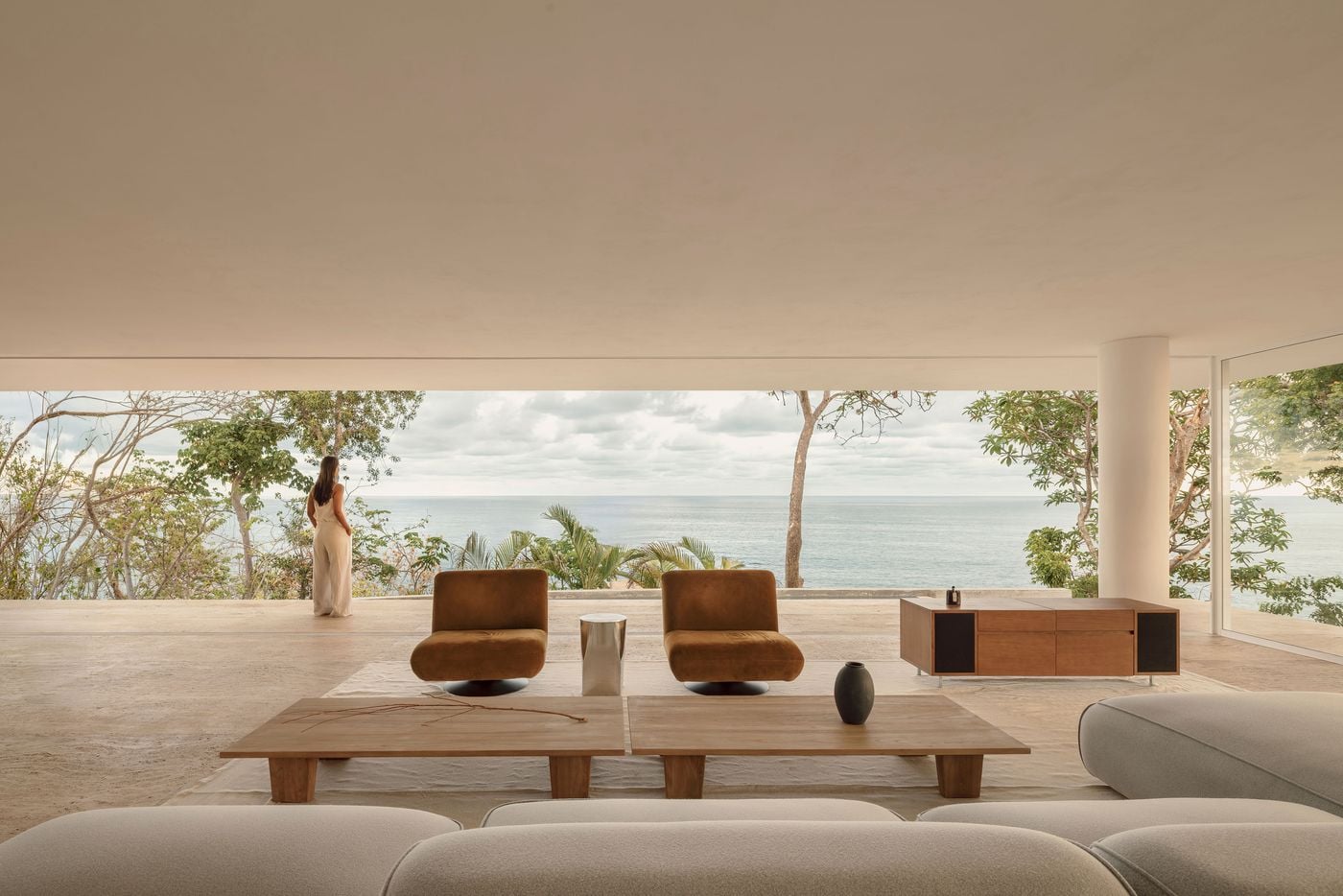 From behind a grey bouclé sofa, the living area unfolds towards an uninterrupted Pacific horizon, framed by the deep ceiling plane and slender trees. Twin cognac velvet swivel chairs, a polished steel side table and paired low timber tables sit on pale veined paving as a woman pauses at the terrace edge. From behind a grey bouclé sofa, the living area unfolds towards an uninterrupted Pacific horizon, framed by the deep ceiling plane and slender trees. Twin cognac velvet swivel chairs, a polished steel side table and paired low timber tables sit on pale veined paving as a woman pauses at the terrace edge.