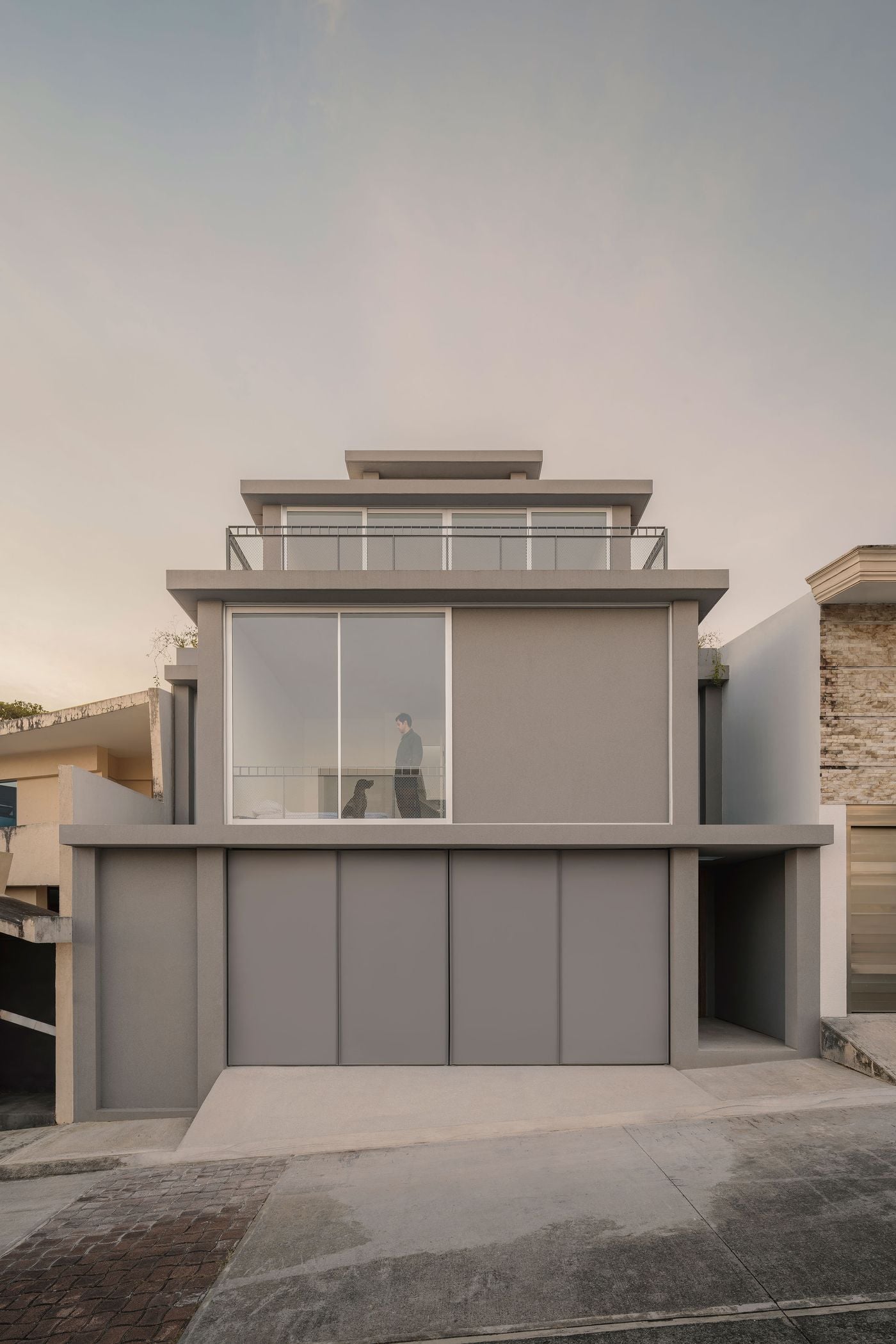 Casa Tlaloc’s street façade presents a restrained composition of stacked grey volumes, broad glazing and projecting horizontal slabs. A large upper-floor window reveals an occupant and dog inside, while metal-mesh balustrades add delicate graphic detail to the otherwise austere elevation above the flush-panelled garage. Casa Tlaloc’s street façade presents a restrained composition of stacked grey volumes, broad glazing and projecting horizontal slabs. A large upper-floor window reveals an occupant and dog inside, while metal-mesh balustrades add delicate graphic detail to the otherwise austere elevation above the flush-panelled garage.