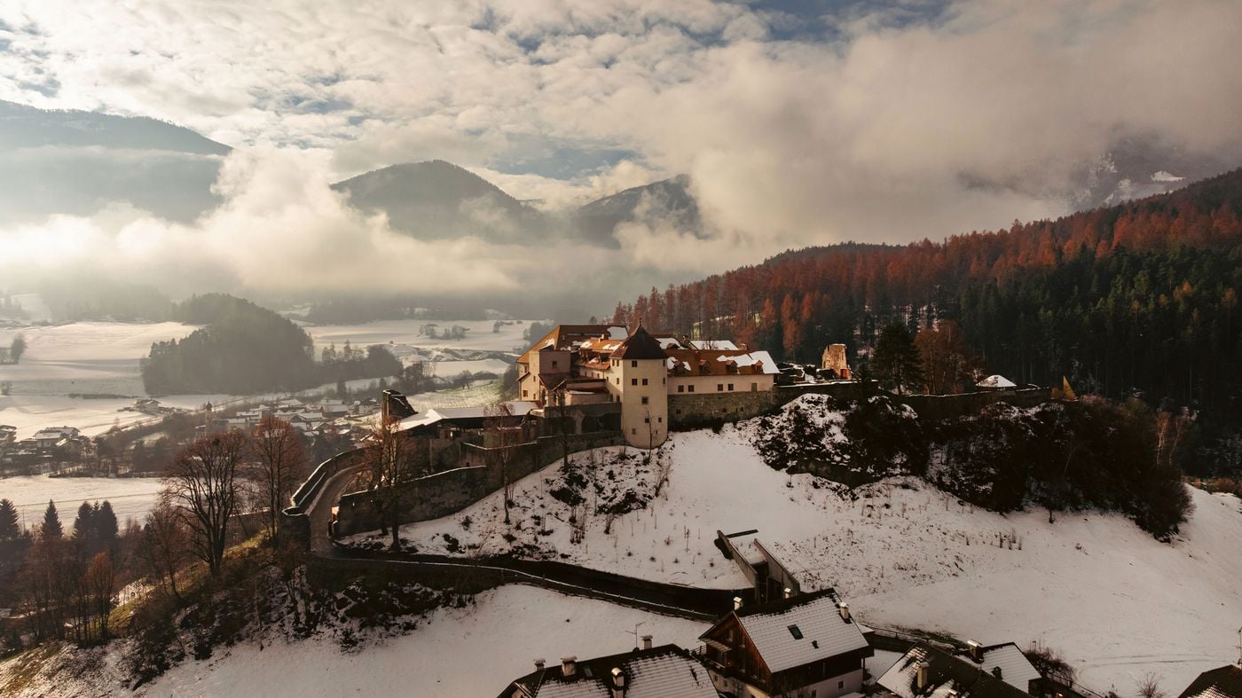 An aerial winter view of Castel Badia shows the castle on its rocky outcrop above the Puster Valley. Pale rendered wings, a round tower and terracotta roofs sit within crenellated ring walls, flanked by russet larch forest. Low cloud drifts over snow-covered fields and the village below. An aerial winter view of Castel Badia shows the castle on its rocky outcrop above the Puster Valley. Pale rendered wings, a round tower and terracotta roofs sit within crenellated ring walls, flanked by russet larch forest. Low cloud drifts over snow-covered fields and the village below.