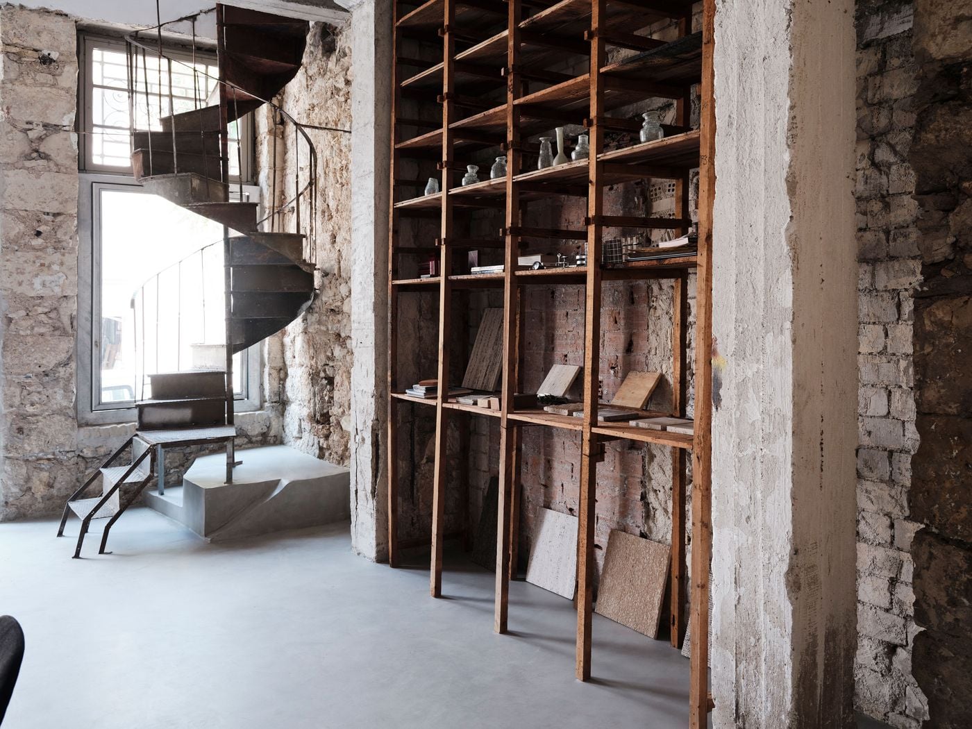 A rusted spiral staircase and tall timber shelving inhabit the exposed masonry shell of MPNYC’s Piraeus studio. Stone, brick, weathered wood and oxidised metal retain their patina against the seamless grey floor, exemplifying the renovation’s approach of incorporating traces of successive occupations rather than erasing them. A rusted spiral staircase and tall timber shelving inhabit the exposed masonry shell of MPNYC’s Piraeus studio. Stone, brick, weathered wood and oxidised metal retain their patina against the seamless grey floor, exemplifying the renovation’s approach of incorporating traces of successive occupations rather than erasing them.