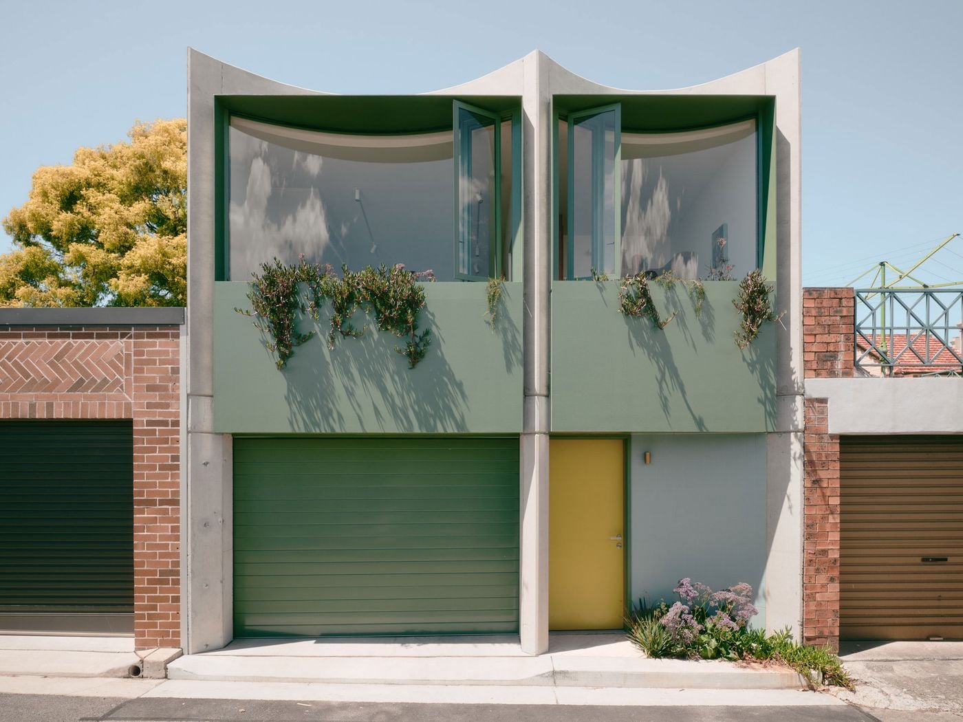Carter Williamson Architects’ Denj Lane presents a playful domestic face to a typically utilitarian Sydney laneway. Twin concave concrete bays frame sage-green glazing and planter boxes, while matching garage doors and a mustard-yellow entrance punctuate the façade, combining sculptural geometry, colour and greenery to signal occupation and care. Carter Williamson Architects’ Denj Lane presents a playful domestic face to a typically utilitarian Sydney laneway. Twin concave concrete bays frame sage-green glazing and planter boxes, while matching garage doors and a mustard-yellow entrance punctuate the façade, combining sculptural geometry, colour and greenery to signal occupation and care.