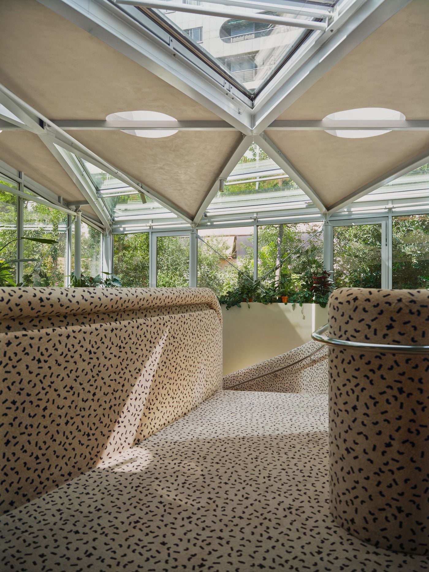 A broad view of the Ken Club’s upholstered staircase reveals its dialogue with the glass-enclosed patio above. Flecked cream surfaces flow continuously across steps, balustrades and landings, while circular roof openings and abundant planting reinforce the project’s recurring interplay of rounded forms, pattern and daylight. A broad view of the Ken Club’s upholstered staircase reveals its dialogue with the glass-enclosed patio above. Flecked cream surfaces flow continuously across steps, balustrades and landings, while circular roof openings and abundant planting reinforce the project’s recurring interplay of rounded forms, pattern and daylight.