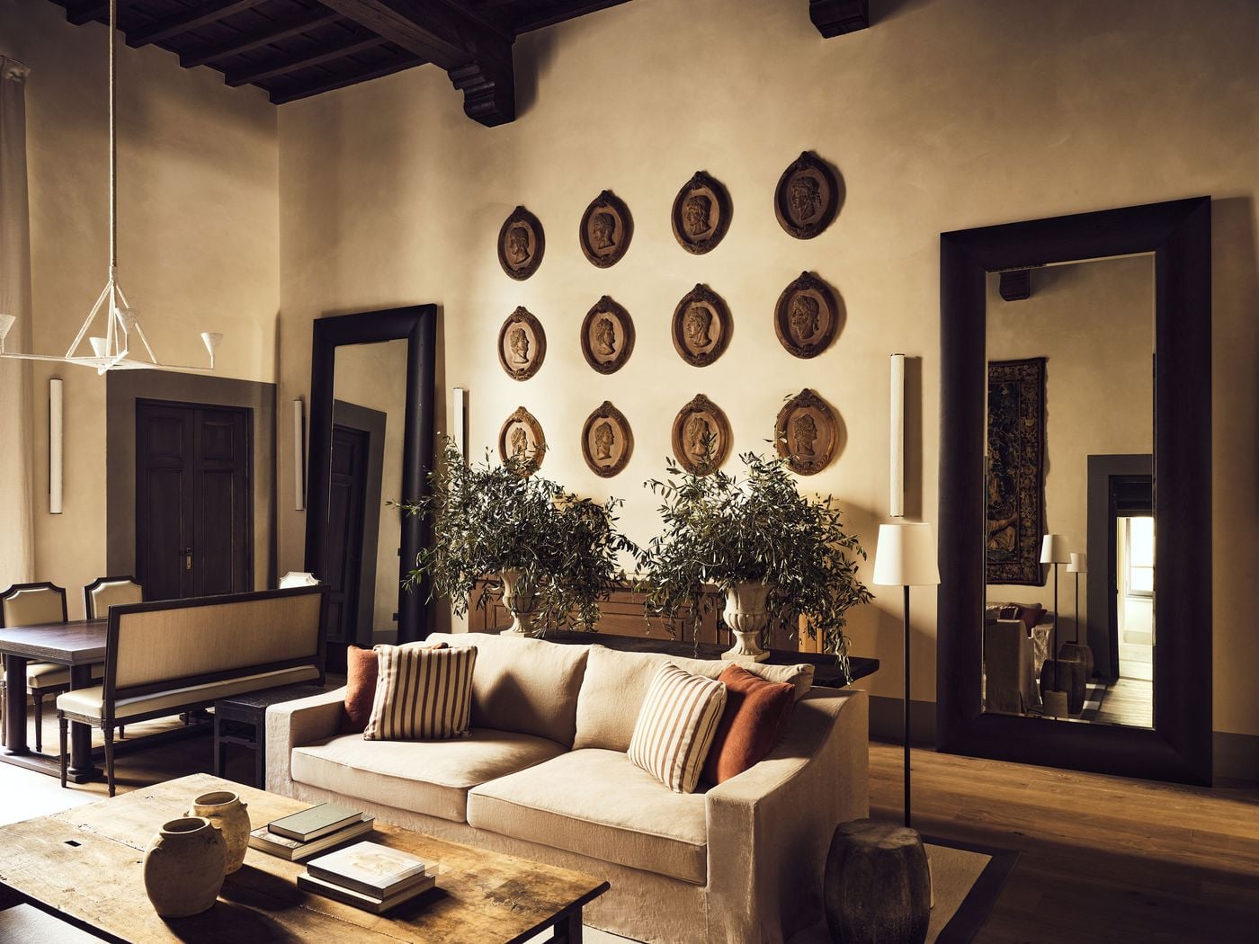 A high-ceilinged living room at La Réserve Firenze, where a grid of twelve classical profile medallions rises above a linen sofa flanked by urns of olive branches. Dark timber beams, two oversized dark-framed mirrors and a sculptural white chandelier give the softly plastered walls a quietly theatrical edge. A high-ceilinged living room at La Réserve Firenze, where a grid of twelve classical profile medallions rises above a linen sofa flanked by urns of olive branches. Dark timber beams, two oversized dark-framed mirrors and a sculptural white chandelier give the softly plastered walls a quietly theatrical edge.