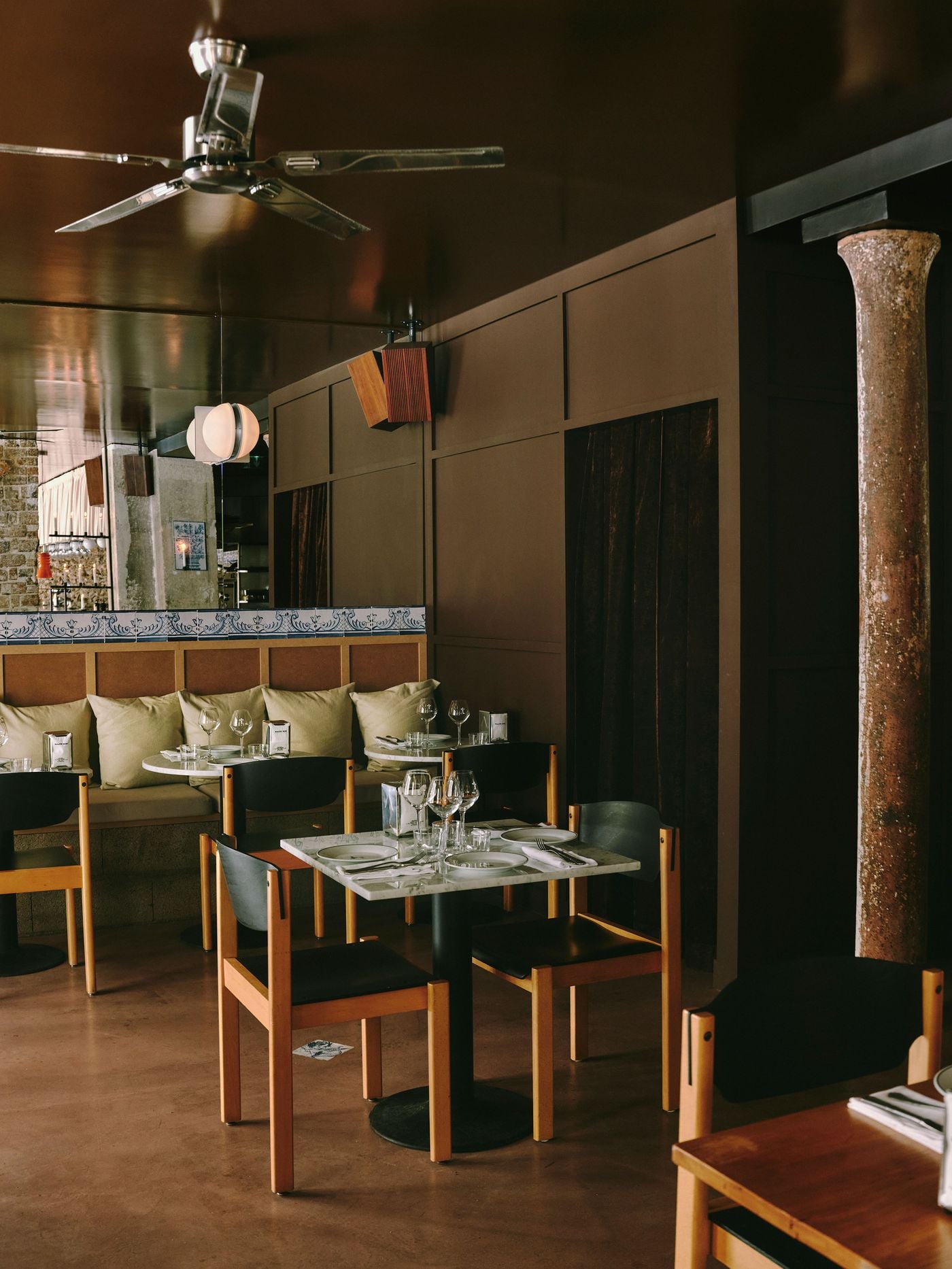 A dining corner at SaltoBar in Paris, where a weathered cast-iron column stands against chocolate-brown panelled walls and velvet-curtained alcoves. Beneath a lacquered ceiling and a steel ceiling fan, black-backed vintage chairs with honey-toned frames gather around marble tables. A frieze of blue-and-white azulejos crowns an MDF-panelled banquette. A dining corner at SaltoBar in Paris, where a weathered cast-iron column stands against chocolate-brown panelled walls and velvet-curtained alcoves. Beneath a lacquered ceiling and a steel ceiling fan, black-backed vintage chairs with honey-toned frames gather around marble tables. A frieze of blue-and-white azulejos crowns an MDF-panelled banquette.