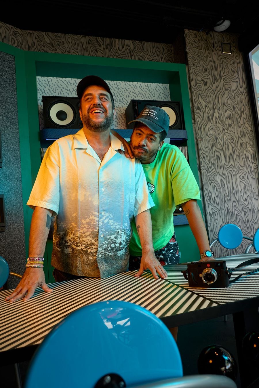 The two founders of Seth & Manni's pose at a striped bistro table, one resting his chin on the other's shoulder. Behind them, a teal-lacquered alcove holds speakers and stacked records against Memphis-patterned backing, framed by grey wood-grain veneer. In the foreground, the glossy blue disc of a First chair looms large. The two founders of Seth & Manni's pose at a striped bistro table, one resting his chin on the other's shoulder. Behind them, a teal-lacquered alcove holds speakers and stacked records against Memphis-patterned backing, framed by grey wood-grain veneer. In the foreground, the glossy blue disc of a First chair looms large.