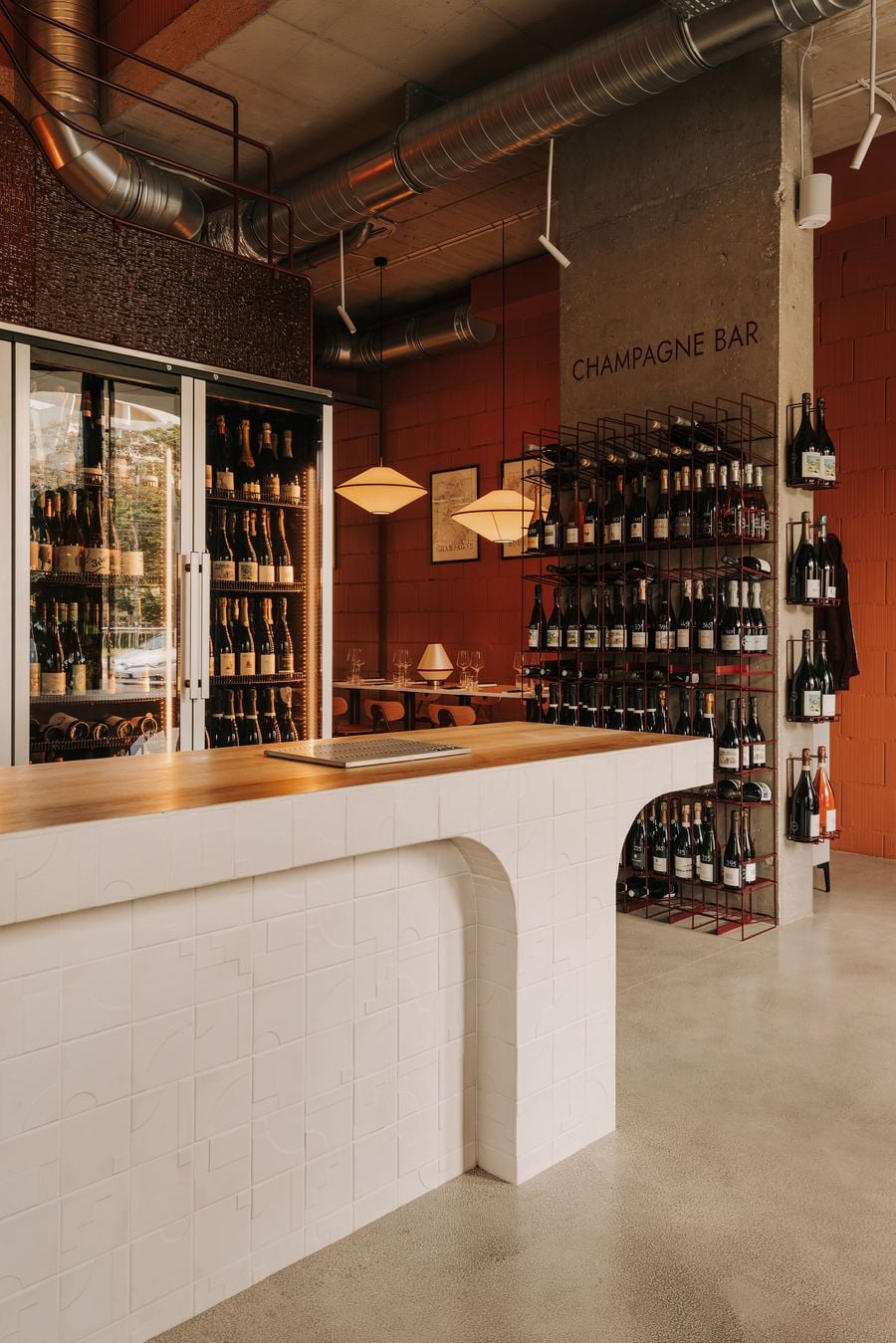 The ground-floor champagne bar at WIN wine bar in Warsaw, designed by Znamy się, juxtaposes a sculptural white-tiled counter with raw concrete, terracotta-coloured masonry and exposed ventilation ducts. Burgundy metal wine racks introduce geometric precision, while warm timber surfaces and softly glowing pendant lights temper the industrial aesthetic. The ground-floor champagne bar at WIN wine bar in Warsaw, designed by Znamy się, juxtaposes a sculptural white-tiled counter with raw concrete, terracotta-coloured masonry and exposed ventilation ducts. Burgundy metal wine racks introduce geometric precision, while warm timber surfaces and softly glowing pendant lights temper the industrial aesthetic.