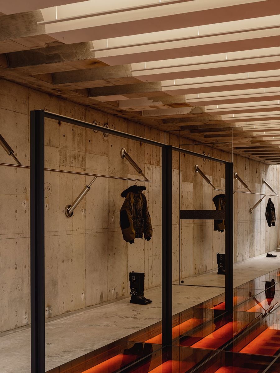 The basement adopts a darker, more linear character, with garments displayed against bare concrete walls beneath a rhythmic ceiling of illuminated white slats. Black metal frames and reflective glazing border the central void, through which the red-lit floor below glows upward, lending the industrial space a theatrical edge.