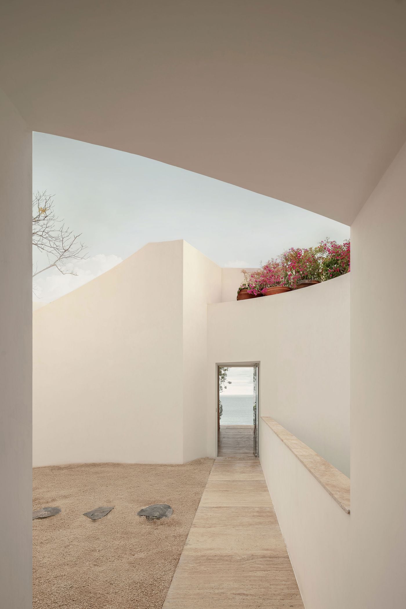 From a shaded threshold, a pale paved walkway runs alongside a gravel court set with dark stones towards a narrow doorway that frames a sliver of sea. Above, white walls step against the sky, crowned by terracotta pots spilling over with pink blossoms. From a shaded threshold, a pale paved walkway runs alongside a gravel court set with dark stones towards a narrow doorway that frames a sliver of sea. Above, white walls step against the sky, crowned by terracotta pots spilling over with pink blossoms.