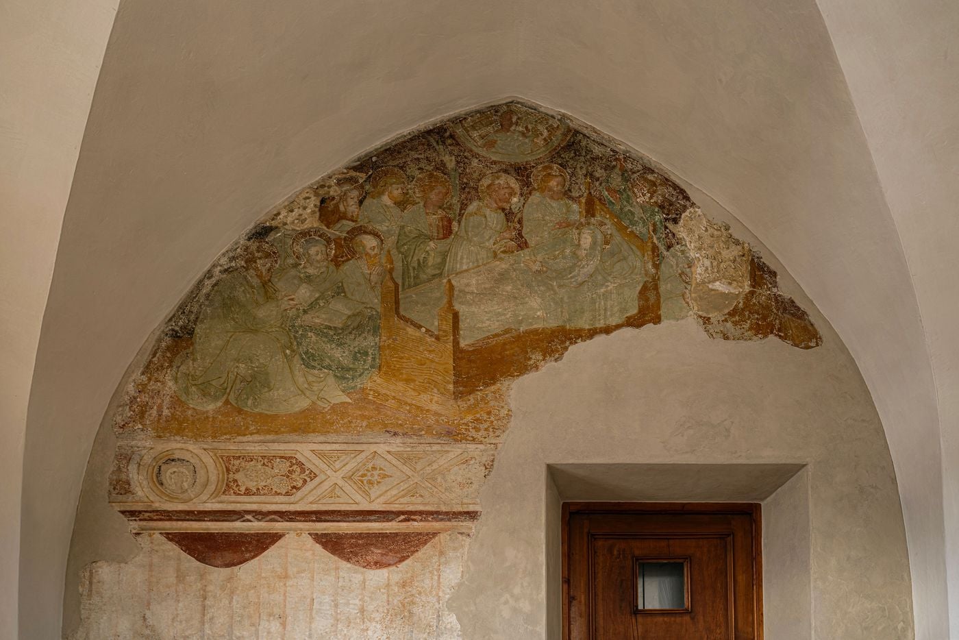 A fragmentary medieval fresco fills a pointed vault at Castel Badia, apparently depicting the Dormition of the Virgin. Haloed apostles in muted greens and ochres gather around Mary's bier beneath a painted geometric frieze and draped border. The surrounding fresh lime plaster frames the surviving pigment like an archaeological find. A fragmentary medieval fresco fills a pointed vault at Castel Badia, apparently depicting the Dormition of the Virgin. Haloed apostles in muted greens and ochres gather around Mary's bier beneath a painted geometric frieze and draped border. The surrounding fresh lime plaster frames the surviving pigment like an archaeological find.