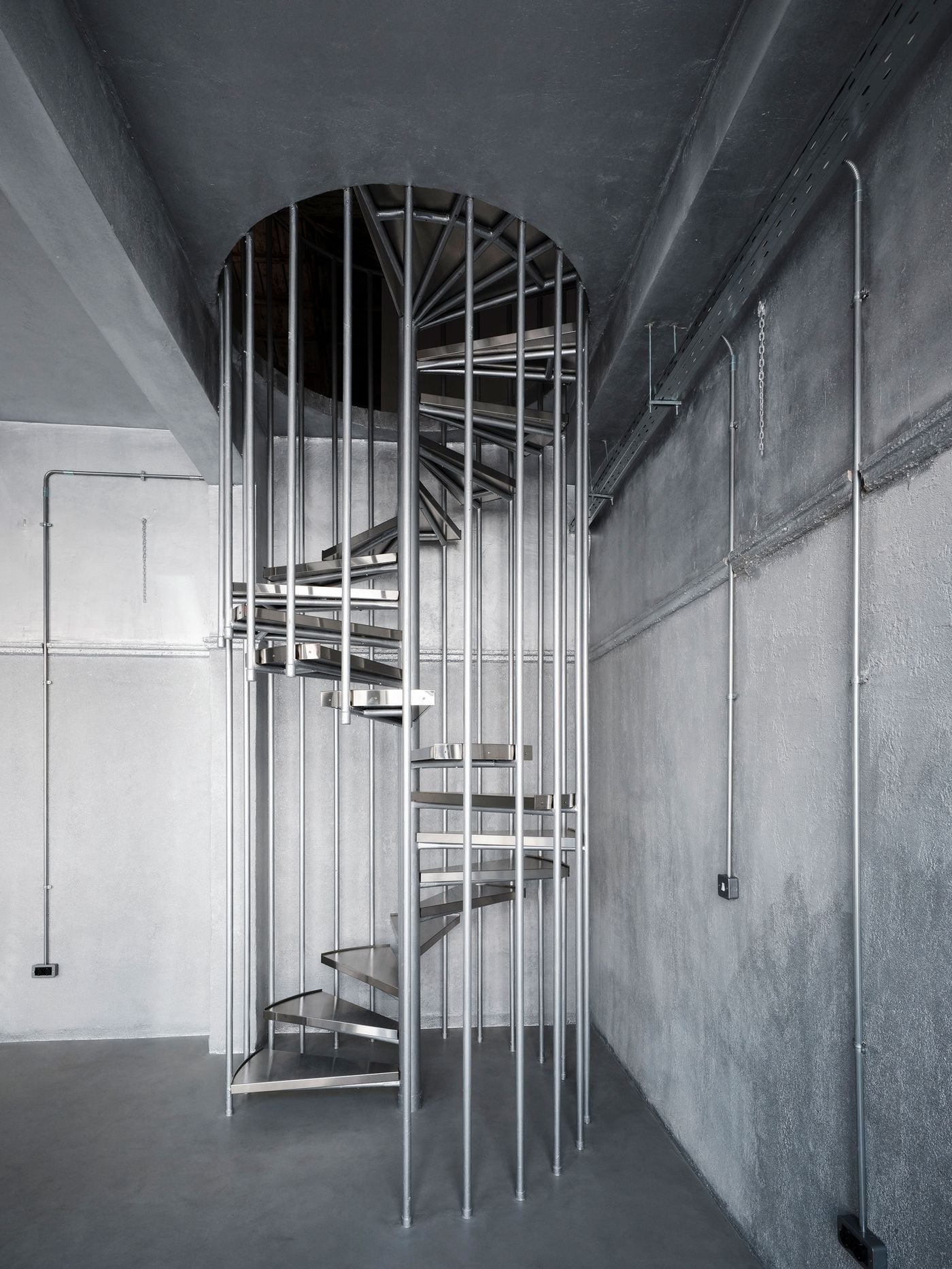 A new metal spiral staircase rises through the monochromatic gallery, surrounded by slender vertical rods that give the structure an almost cage-like appearance. Its metallic surfaces echo the silvery finish enveloping the walls and ceiling, while exposed conduits and cable trays reinforce the space’s industrial character. A new metal spiral staircase rises through the monochromatic gallery, surrounded by slender vertical rods that give the structure an almost cage-like appearance. Its metallic surfaces echo the silvery finish enveloping the walls and ceiling, while exposed conduits and cable trays reinforce the space’s industrial character.