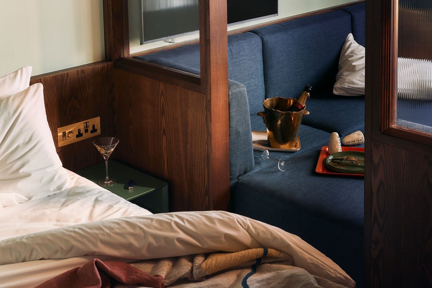 A deliberately lived-in vignette captures the relationship between the bed and adjoining blue sofa pit, separated by dark walnut framing and reeded glass. Rumpled bedding, martini glasses and a champagne bucket lend the scene an after-hours mood, echoing the playful 1960s hedonism underpinning Holloway Li’s concept. A deliberately lived-in vignette captures the relationship between the bed and adjoining blue sofa pit, separated by dark walnut framing and reeded glass. Rumpled bedding, martini glasses and a champagne bucket lend the scene an after-hours mood, echoing the playful 1960s hedonism underpinning Holloway Li’s concept.