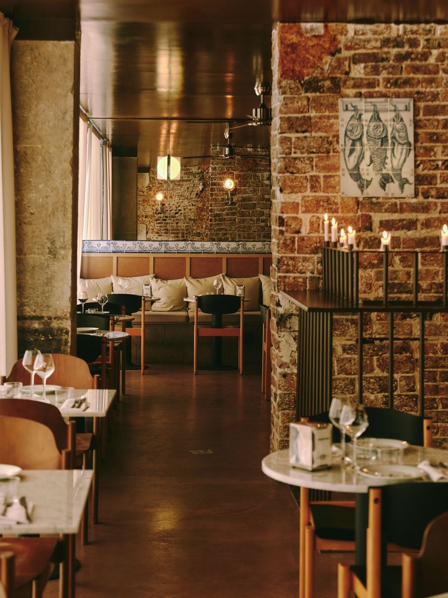 A portrait-format view down the dining room at SaltoBar in Paris, where a burnished ceiling reflects the warm light. Timber-framed chairs with black backrests face a cushioned banquette crowned by an azulejo border. In the foreground, a fish-motif tile panel and a row of flickering candles animate the exposed brick.