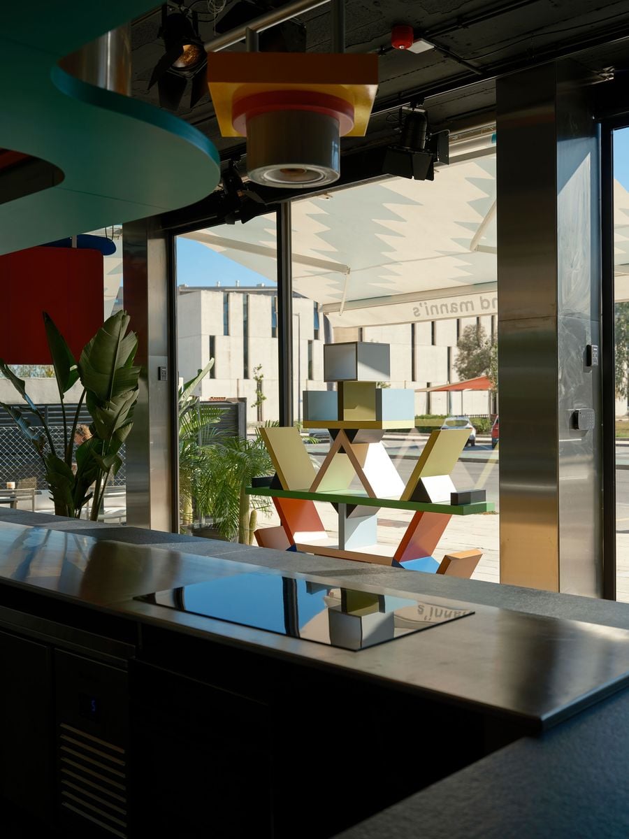 Seen from across the kitchen counter, Ettore Sottsass’ Carlton bookcase is silhouetted against the glazed façade, its colourful geometry mirrored in the black glass of an induction hob. A teal overhang and yellow pendant light frame the upper edge. The zigzag awning and a white modernist building beyond complete the layered composition. Seen from across the kitchen counter, Ettore Sottsass’ Carlton bookcase is silhouetted against the glazed façade, its colourful geometry mirrored in the black glass of an induction hob. A teal overhang and yellow pendant light frame the upper edge. The zigzag awning and a white modernist building beyond complete the layered composition.