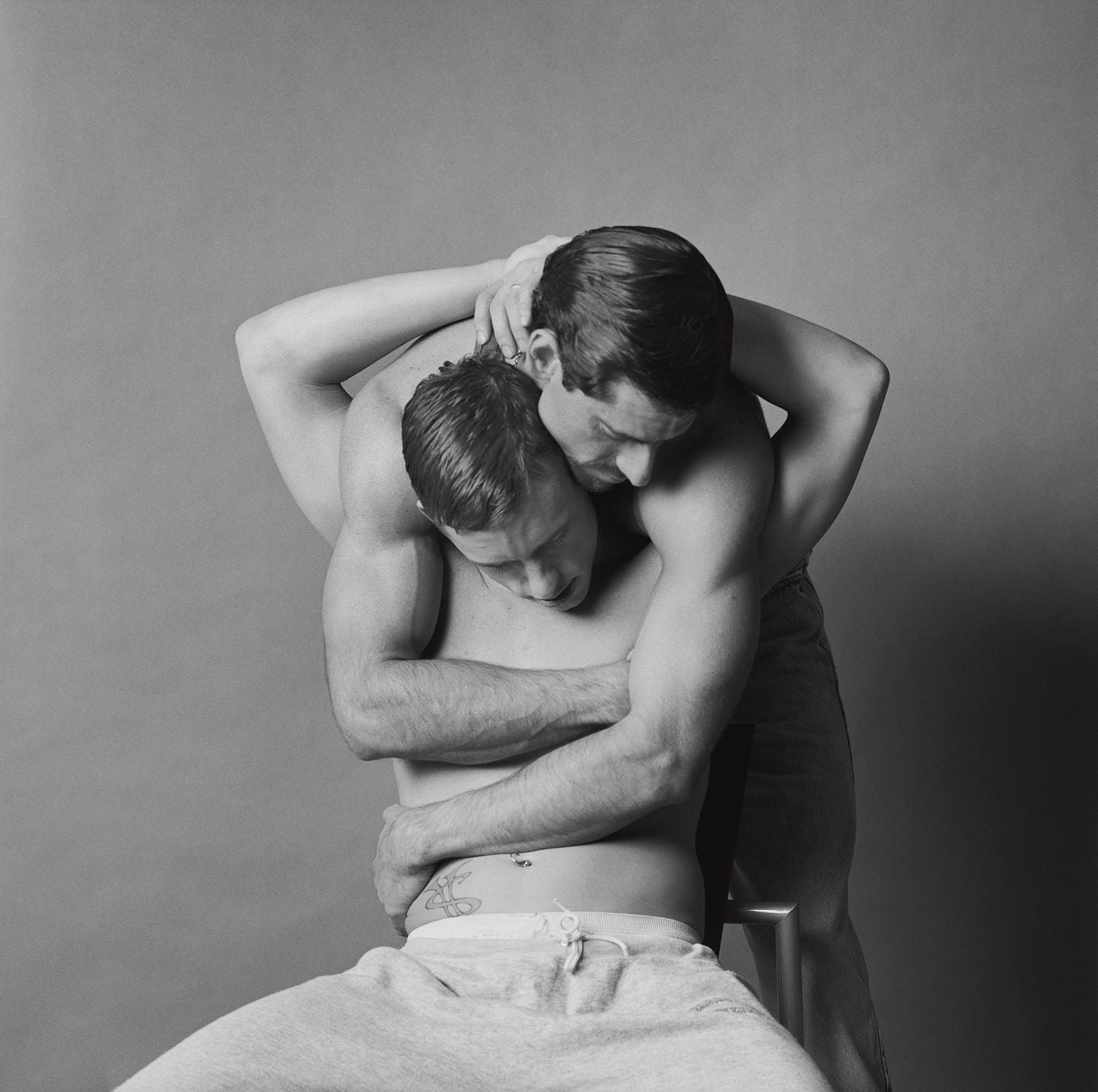 Against a plain studio backdrop, two men fold into a wrestler-like embrace: one leans over from behind, his arms encircling the other's waist, while his seated partner reaches back to cradle his head. A small hip tattoo, a navel piercing and soft grey sweatpants add intimate detail to this tender portrait from Don't Worry Boys. Against a plain studio backdrop, two men fold into a wrestler-like embrace: one leans over from behind, his arms encircling the other's waist, while his seated partner reaches back to cradle his head. A small hip tattoo, a navel piercing and soft grey sweatpants add intimate detail to this tender portrait from Don't Worry Boys.