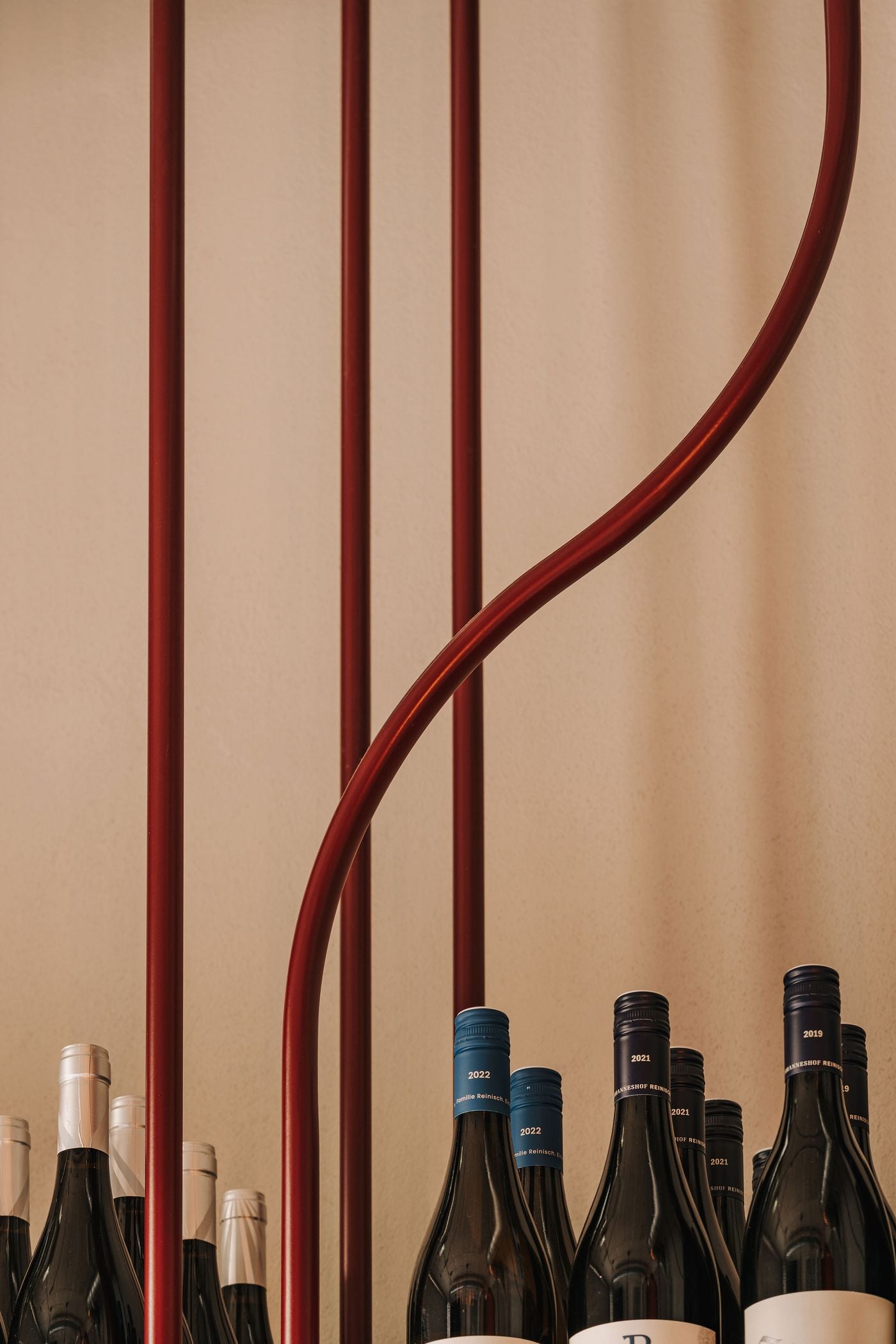 A close-up of WIN's wine shelving highlights the interplay between straight burgundy metal uprights and a sinuous tubular element that curves across the composition. Evoking the movement of climbing vines, the sculptural metalwork introduces an organic counterpoint to the orderly arrangement of wine bottles and the understated beige backdrop. A close-up of WIN's wine shelving highlights the interplay between straight burgundy metal uprights and a sinuous tubular element that curves across the composition. Evoking the movement of climbing vines, the sculptural metalwork introduces an organic counterpoint to the orderly arrangement of wine bottles and the understated beige backdrop.