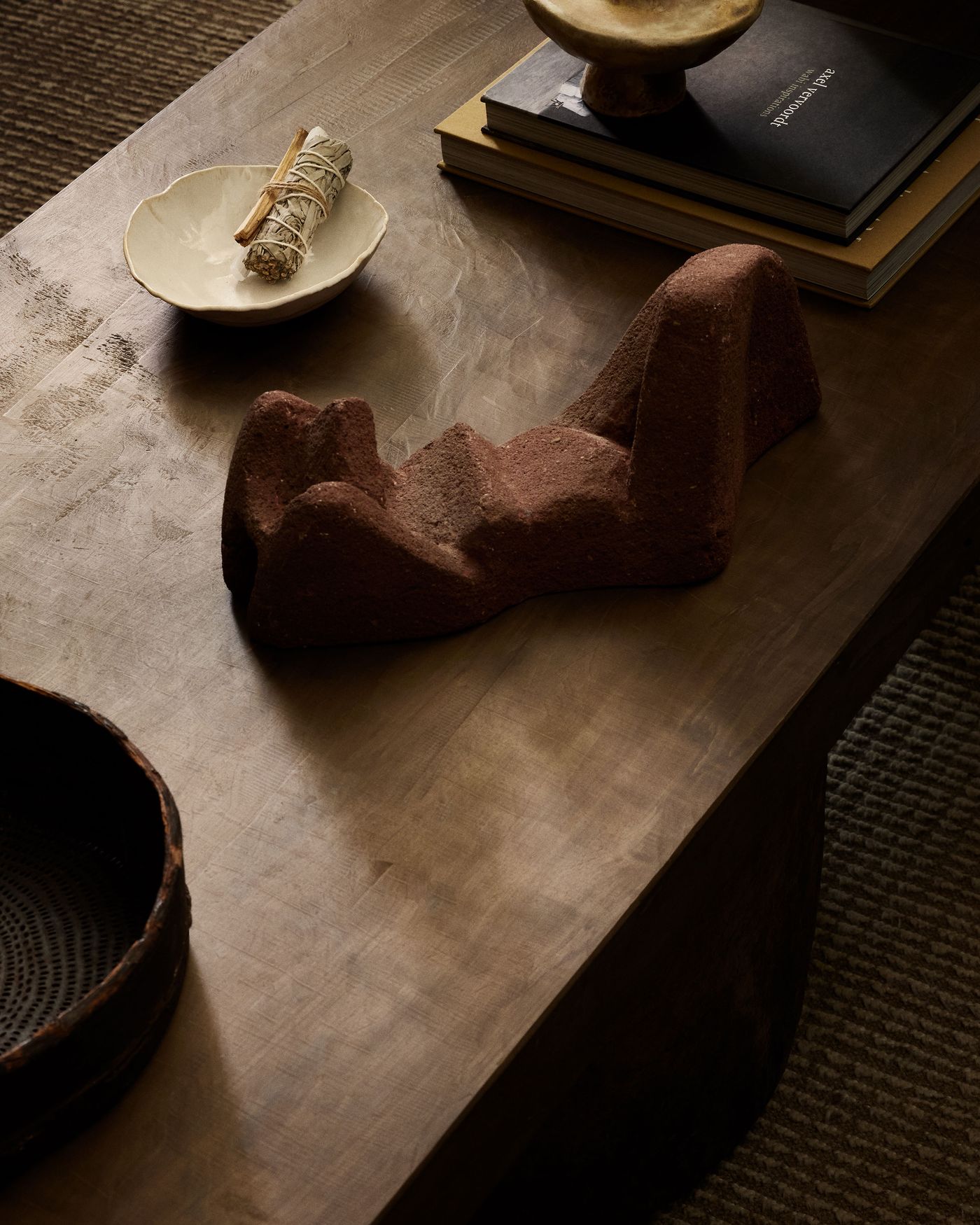 A wooden table displays a sculptural red piece, a white bowl with sage, stacked books, and a small ceramic dish.