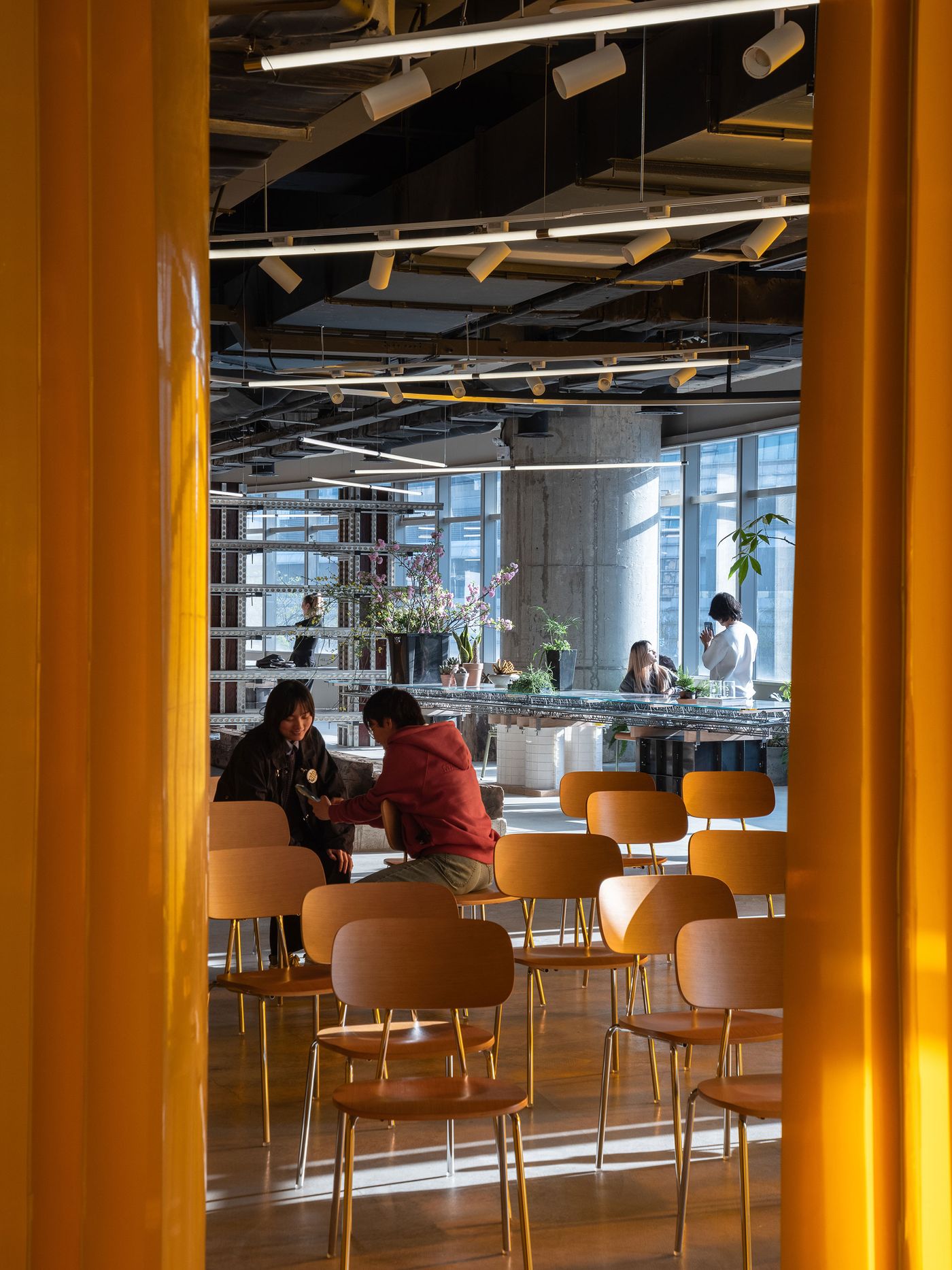 Framed by a striking yellow curtain, this view into 889 GLO Art Space reveals a gathering area with light wooden chairs arranged for an event. Beyond, people mingle in the sunlit general exhibition space, characterized by large windows and the prominent industrial-style shelving, showcasing the venue's versatility and vibrant atmosphere. Framed by a striking yellow curtain, this view into 889 GLO Art Space reveals a gathering area with light wooden chairs arranged for an event. Beyond, people mingle in the sunlit general exhibition space, characterized by large windows and the prominent industrial-style shelving, showcasing the venue's versatility and vibrant atmosphere.