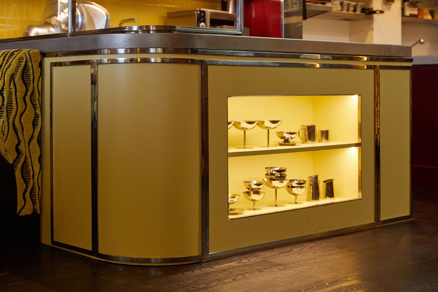 A low-angle shot of a custom-built counter cabinet with a warm, mellow yellow finish and chrome trim. The rounded corner and subtly reflective surface evoke a retro-modernist aesthetic. An integrated, illuminated shelf reveals a curated collection of polished metallic drinking vessels, drawing the eye to the artful display. A low-angle shot of a custom-built counter cabinet with a warm, mellow yellow finish and chrome trim. The rounded corner and subtly reflective surface evoke a retro-modernist aesthetic. An integrated, illuminated shelf reveals a curated collection of polished metallic drinking vessels, drawing the eye to the artful display.