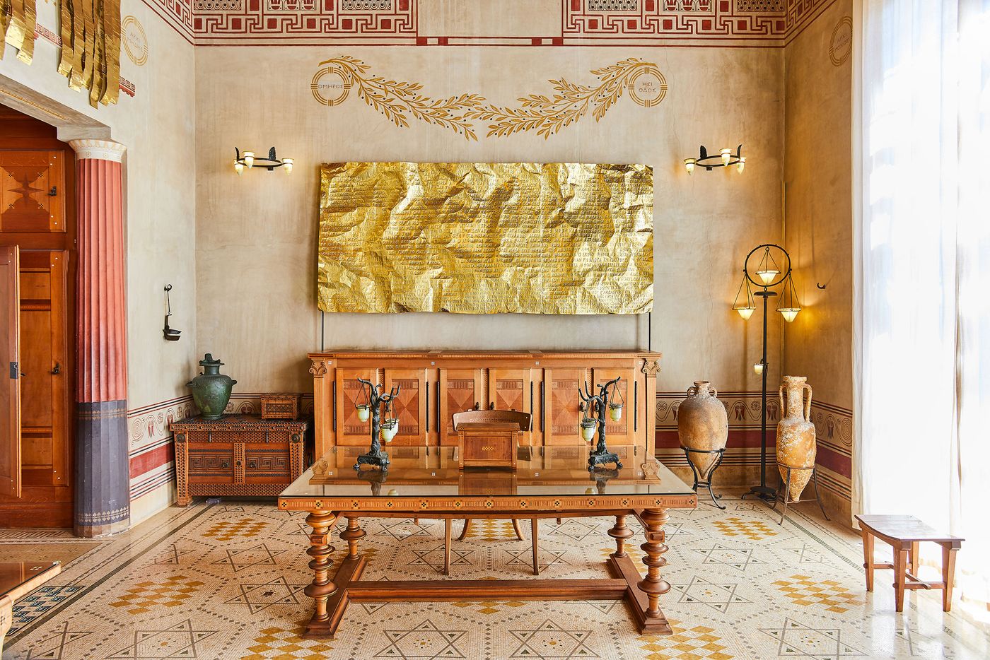 A grand interior room at Villa Kerylos featuring a long wooden console table and a glass-topped dining table with carved wooden legs. Above the console, a large, wrinkled sheet of golden metal with etched text, a modern art piece by Gabriel Leger, hangs against a wall decorated with classical motifs and friezes. A grand interior room at Villa Kerylos featuring a long wooden console table and a glass-topped dining table with carved wooden legs. Above the console, a large, wrinkled sheet of golden metal with etched text, a modern art piece by Gabriel Leger, hangs against a wall decorated with classical motifs and friezes.
