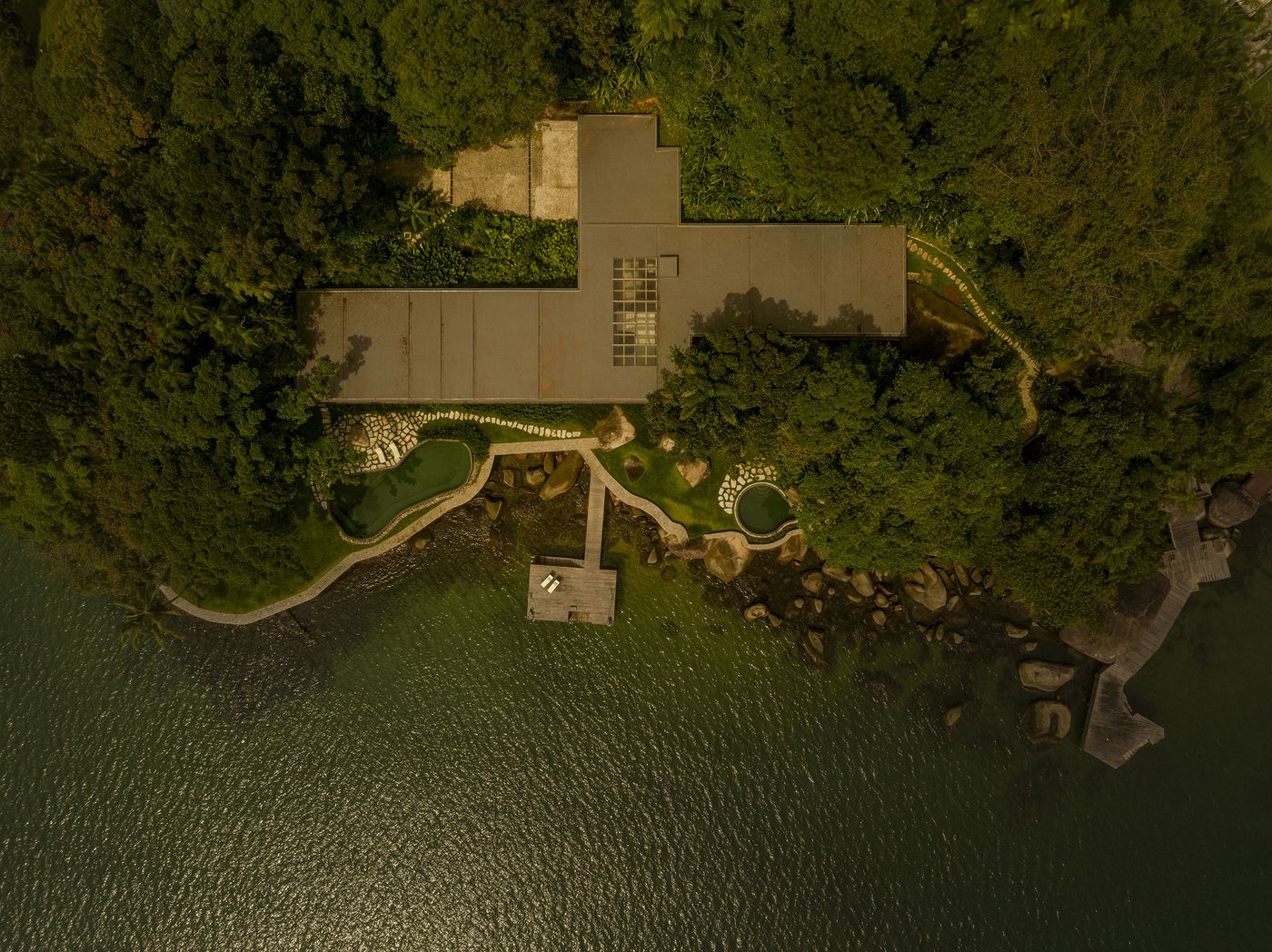 An overhead drone shot reveals the "L" shaped modern layout of the dwelling, discreetly nestled into the natural contours of the lush peninsula. The flat, integrated roofs, natural stone pathways, and organic-shaped pools blend seamlessly with the dense green foliage and the emerald-toned ocean, highlighting a biophilic design. An overhead drone shot reveals the "L" shaped modern layout of the dwelling, discreetly nestled into the natural contours of the lush peninsula. The flat, integrated roofs, natural stone pathways, and organic-shaped pools blend seamlessly with the dense green foliage and the emerald-toned ocean, highlighting a biophilic design.