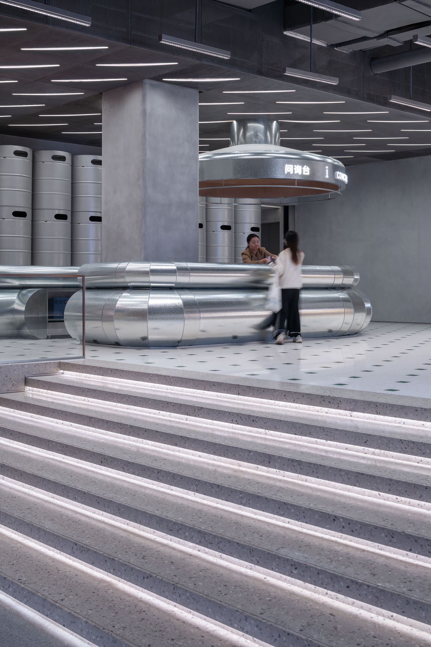 A modern reception area featuring a sleek, metallic counter, illuminated steps, and cylindrical storage units in the background.