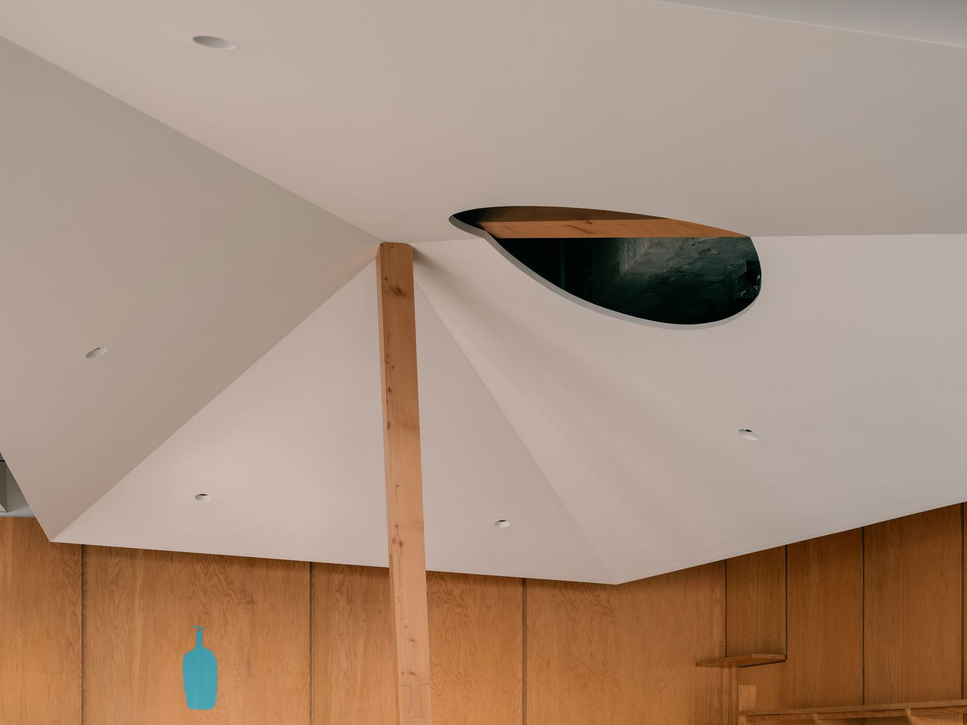 An upward view captures the dynamic geometry of the folded ceiling, anchored by a single timber beam piercing an oval skylight—an architectural gesture that defines the café’s identity.