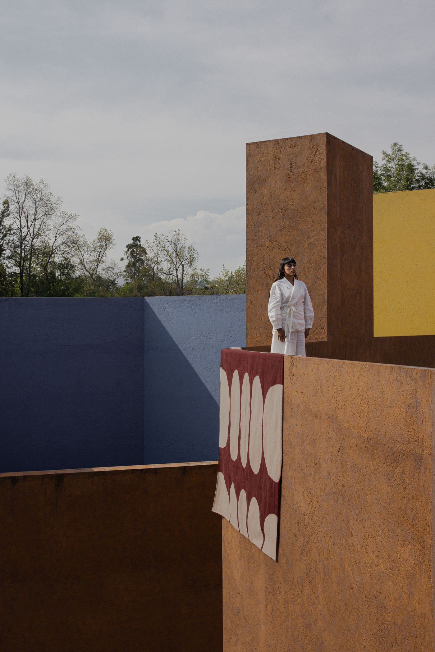 A person in a white outfit stands on a colorful architectural structure with earthy tones and vibrant blue and yellow walls.