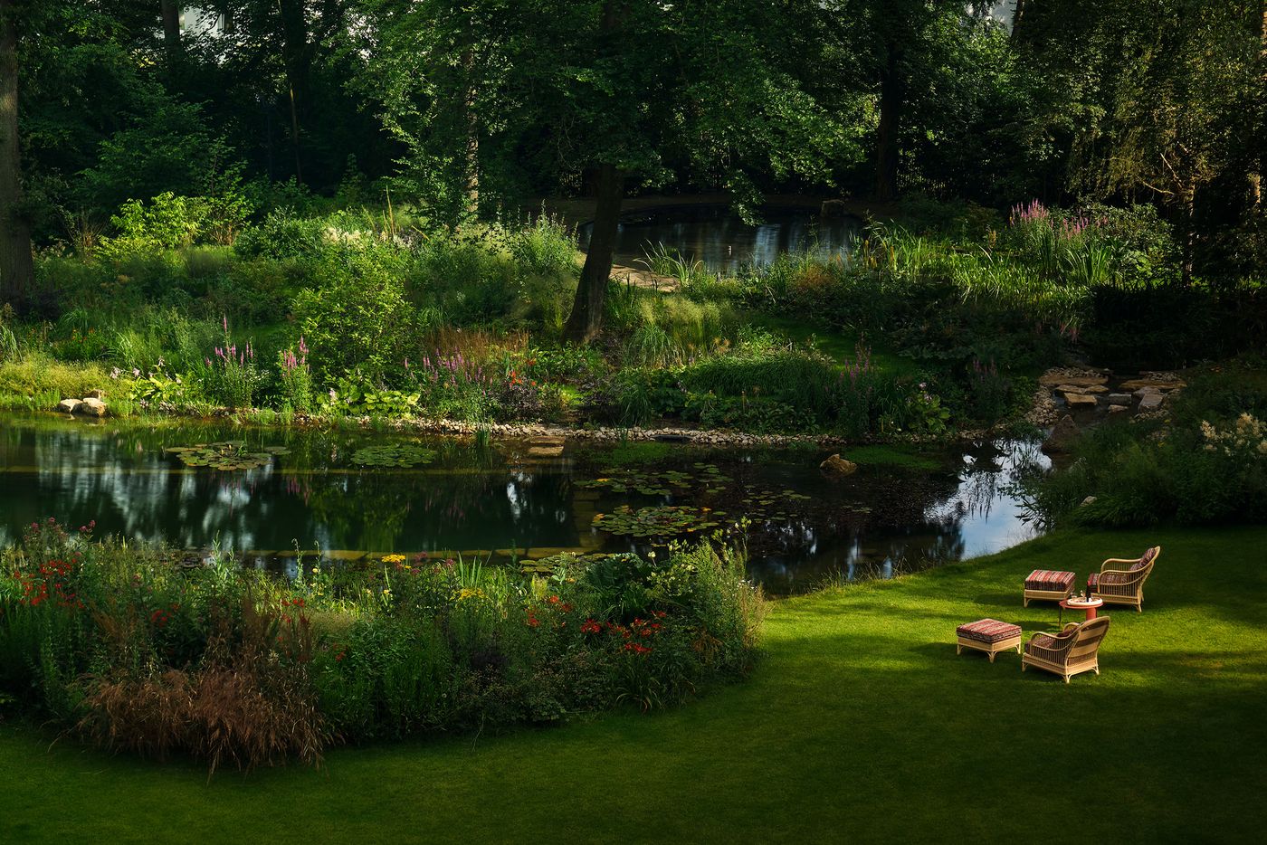 A tranquil garden scene with a pond surrounded by lush greenery, flowers, and two wicker chairs set up for relaxation.