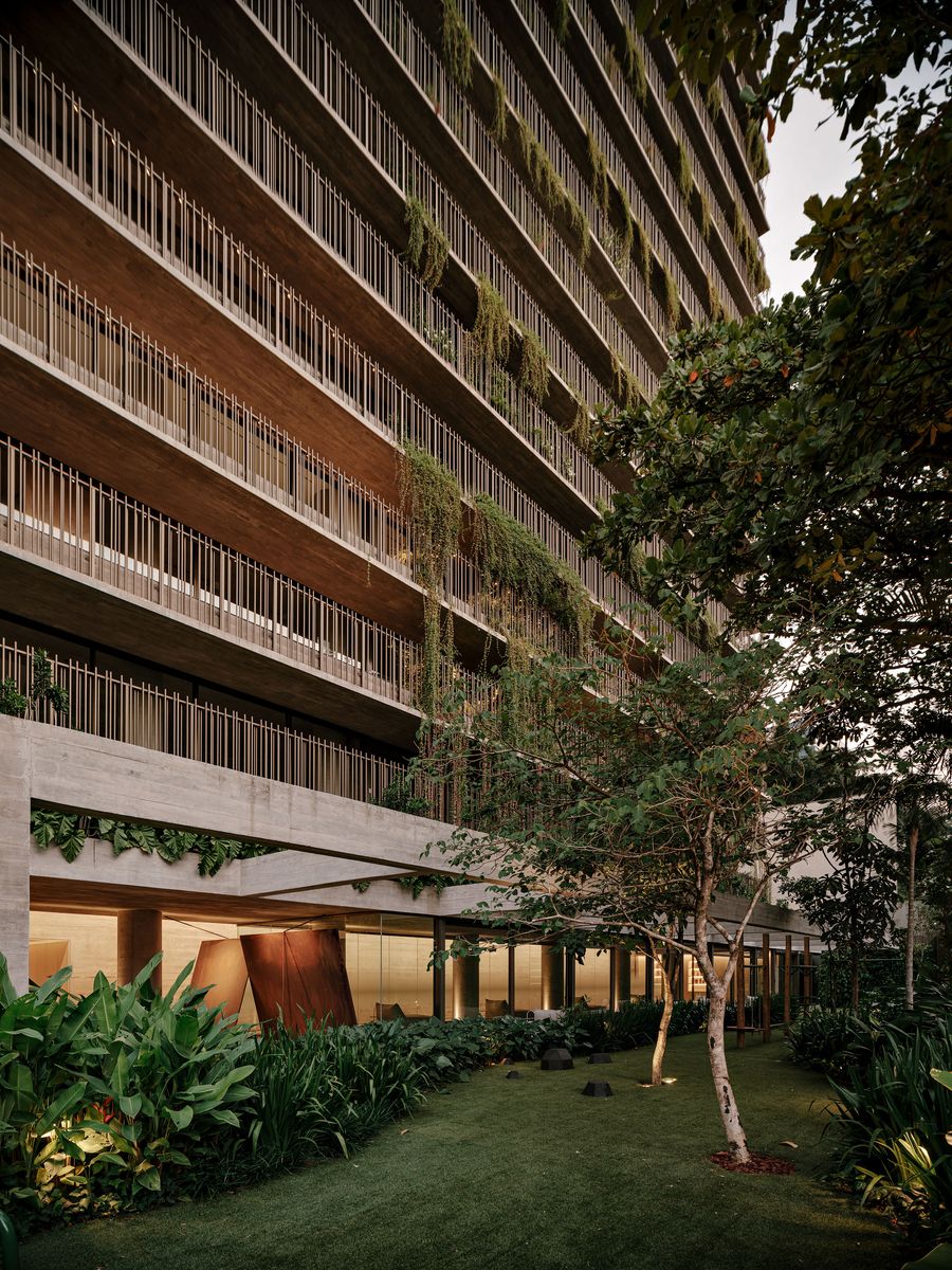 An evening view of the Ibaté building's lower levels, where the concrete structure and extensive greenery are artfully illuminated. Warm interior lights glow through large glass panes, creating a welcoming contrast with the deep shadows of the surrounding garden, highlighting the biophilic brutalist design by Studio Arthur Casas. An evening view of the Ibaté building's lower levels, where the concrete structure and extensive greenery are artfully illuminated. Warm interior lights glow through large glass panes, creating a welcoming contrast with the deep shadows of the surrounding garden, highlighting the biophilic brutalist design by Studio Arthur Casas.