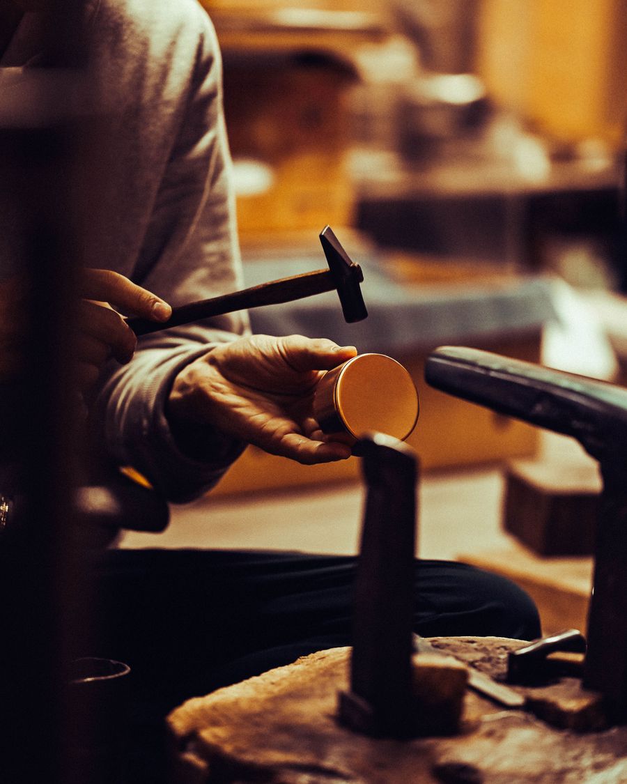 A craftsman holds a round metal piece in one hand while hammering it with a tool, surrounded by a workshop filled with tools. A craftsman holds a round metal piece in one hand while hammering it with a tool, surrounded by a workshop filled with tools.